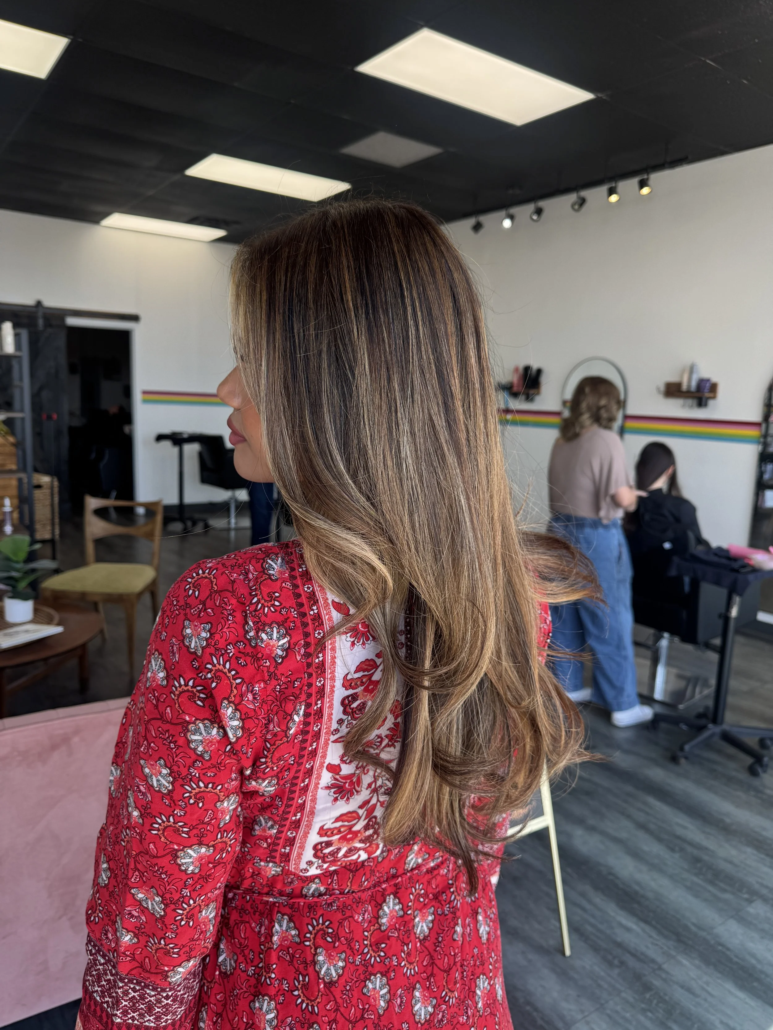 Side profile of a woman with long, wavy, brown hair wearing a red patterned dress inside a salon or studio with a black ceiling and white walls, with other people visible in the background.