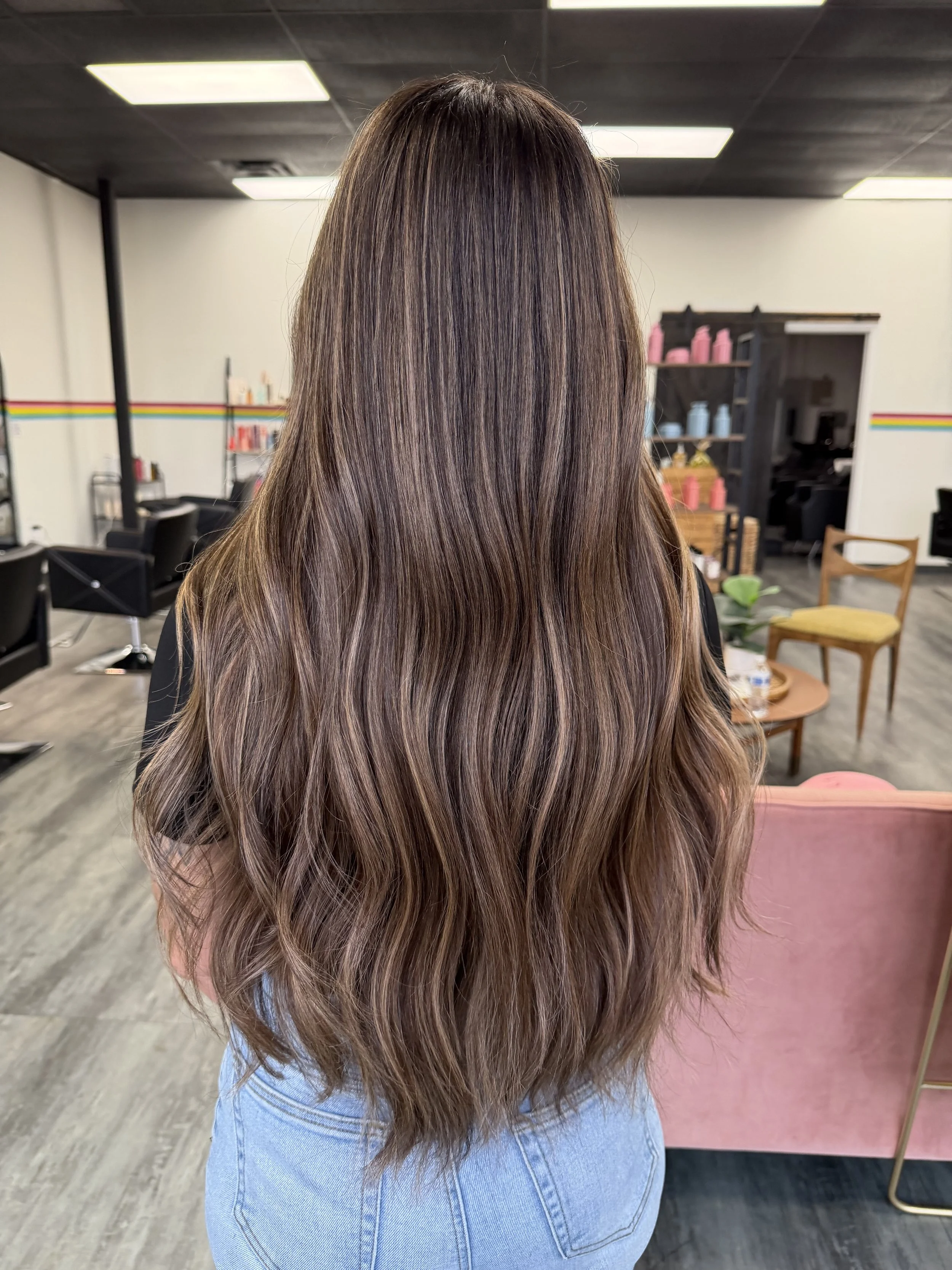 A woman with long, wavy hair with highlights in a salon. She is wearing a black top and blue jeans, facing away from the camera, with salon chairs and shelves in the background.