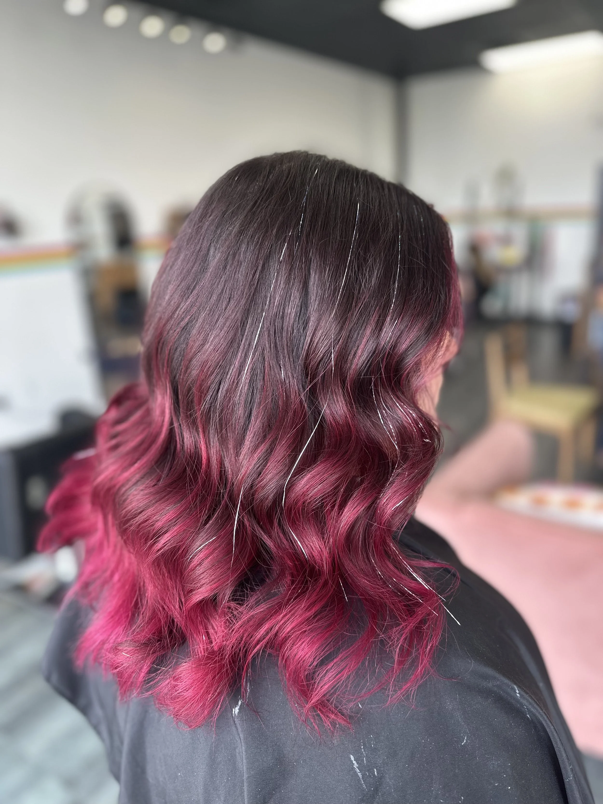 A person with long, wavy hair dyed in shades of black, purple, and pink, sitting in a room with a blurred background of furniture and a table.