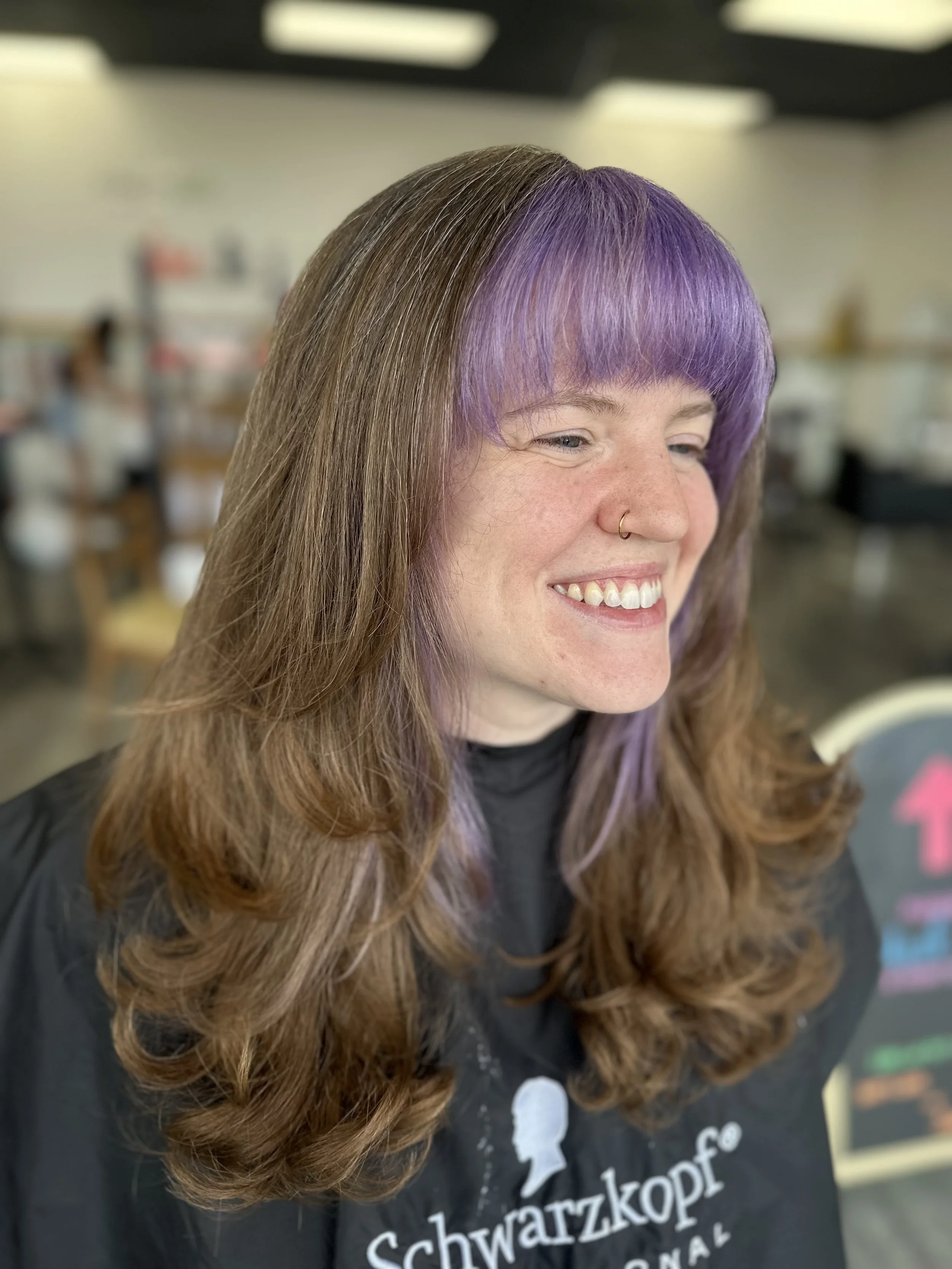 A woman with long brown hair, with a section of her hair dyed purple, smiling. She has a nose ring and is wearing a black shirt with white and pink text.