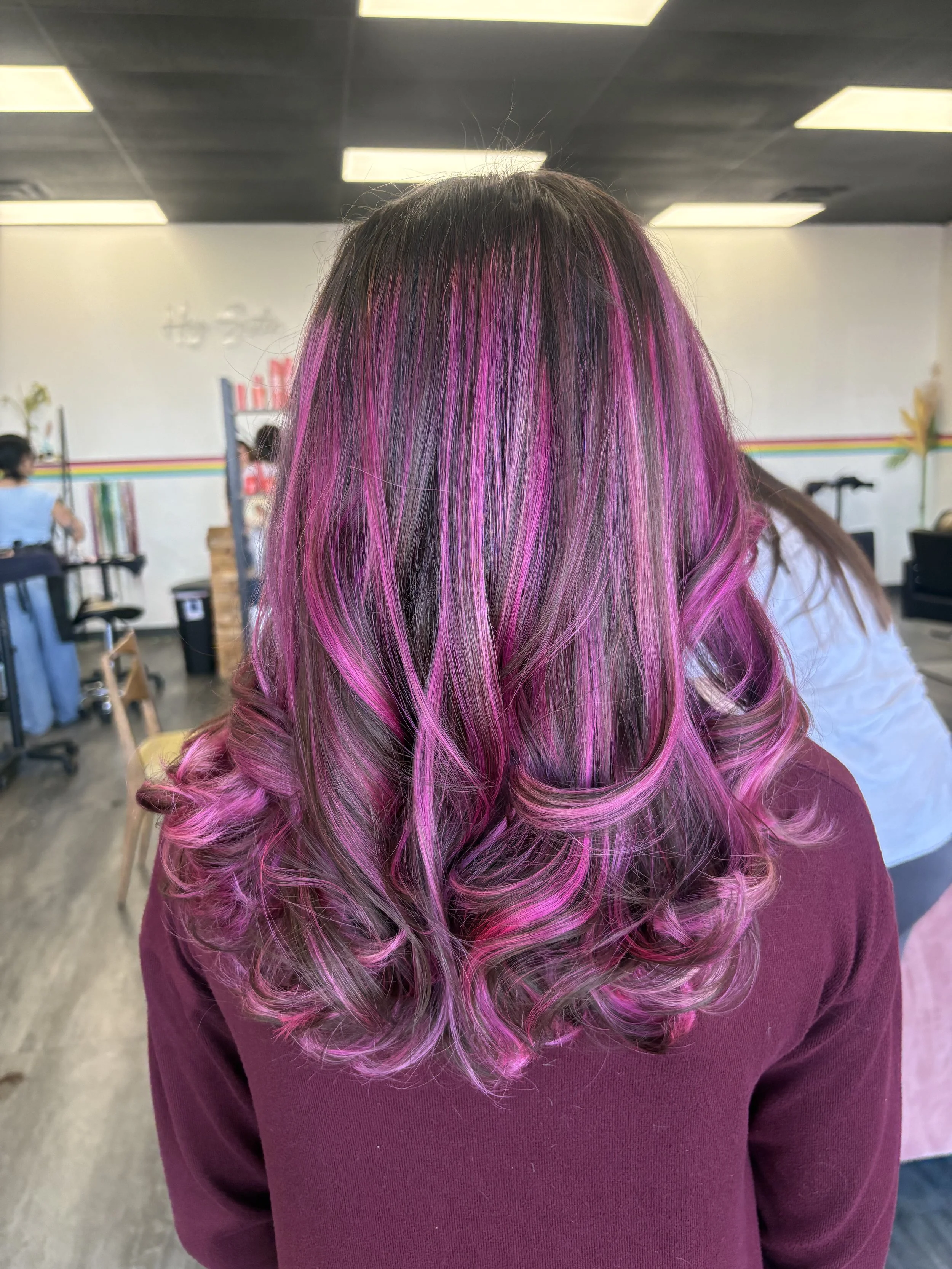 A person with long, curly hair dyed in shades of pink and purple, wearing a maroon shirt, standing indoors with a colorful striped wall and ceiling lights in the background.