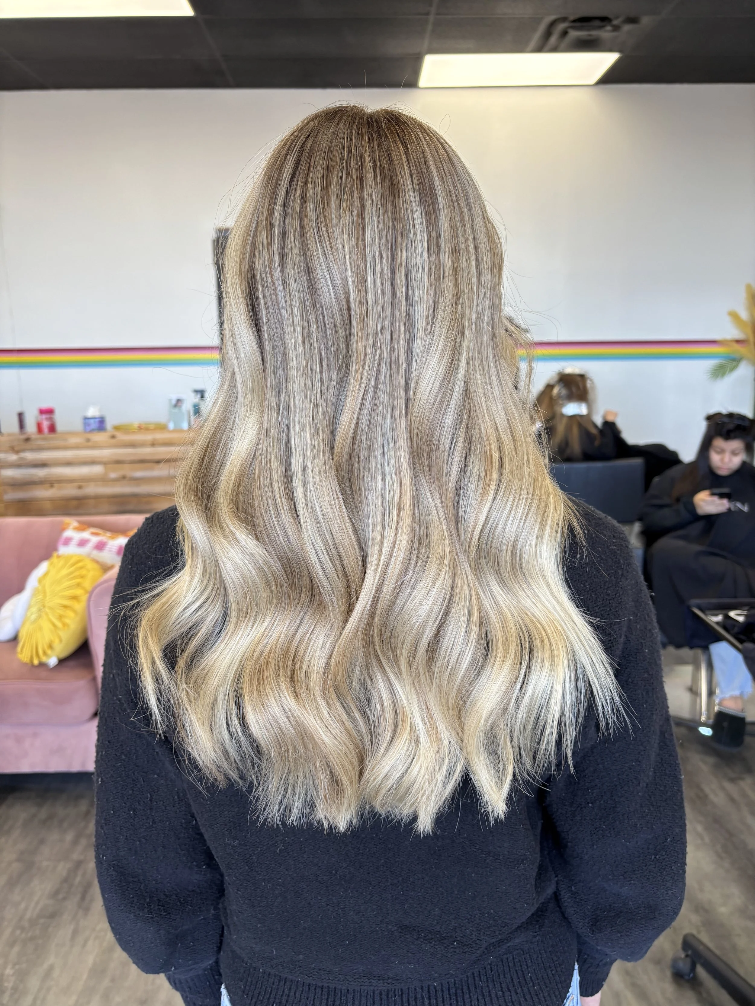 Back view of a woman with long wavy blonde hair with darker roots, wearing a black sweater, in a salon with a colorful rainbow stripe on a white wall and other people in the background.