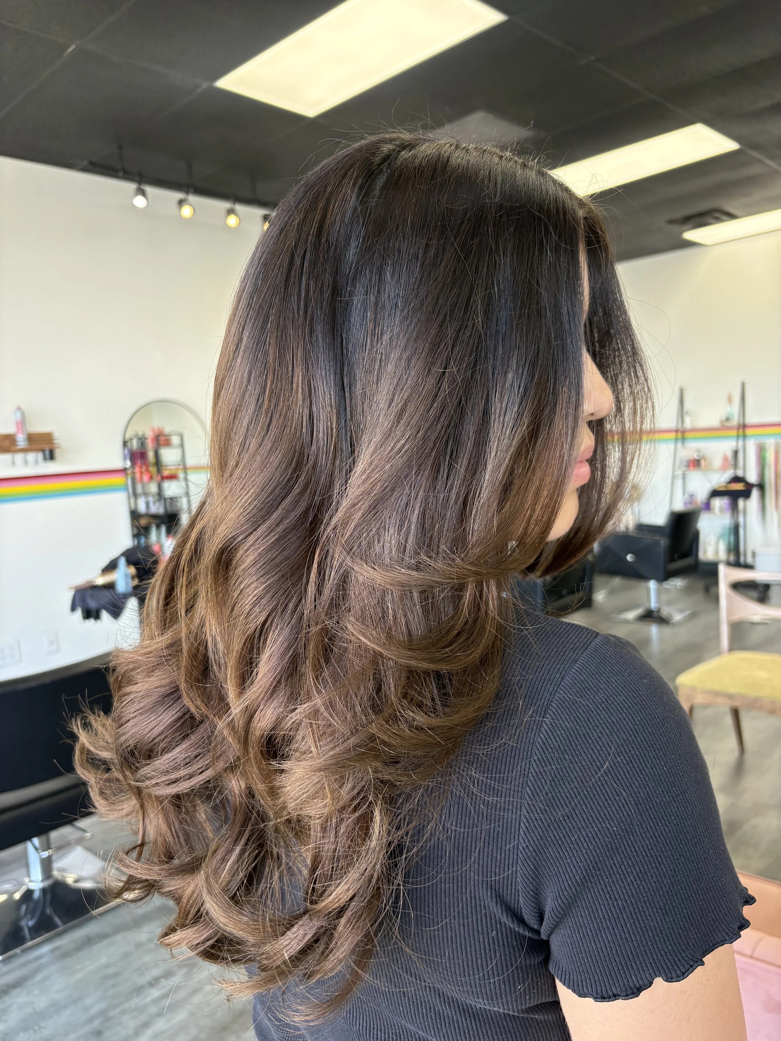 A woman with long, wavy, brown hair with blonde highlights, wearing a black shirt with ruffle sleeves, sitting in a salon or studio with a hair mirror, shelves, and chairs in the background.
