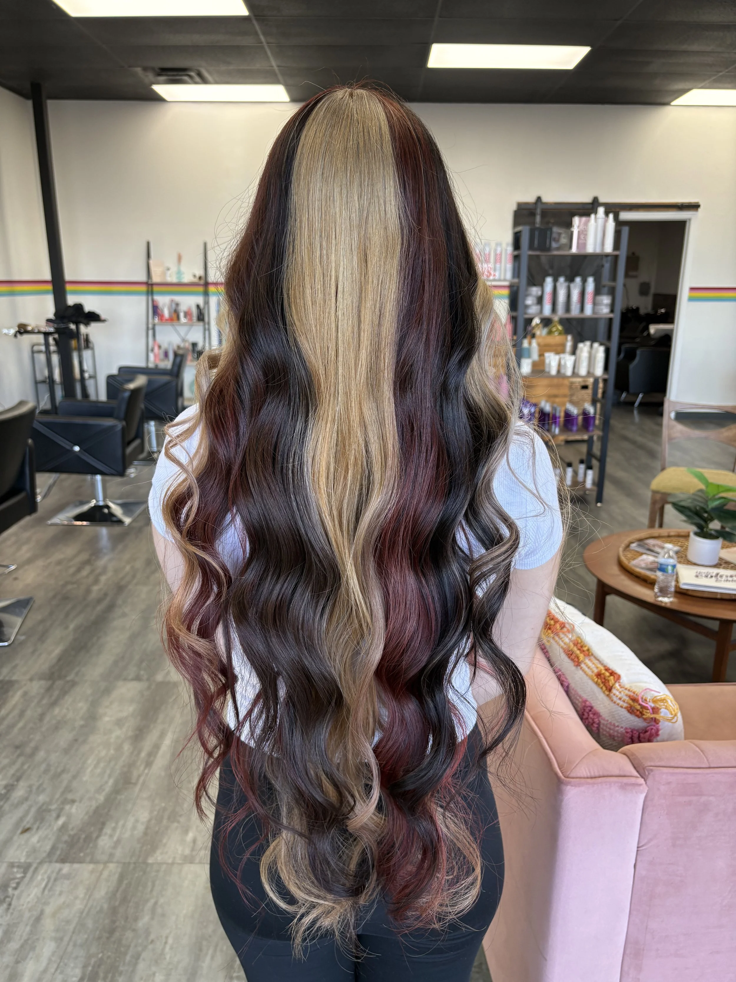 Back view of a woman with long, wavy hair dyed in a blend of blonde, burgundy, and dark brown colors, inside a hair salon.