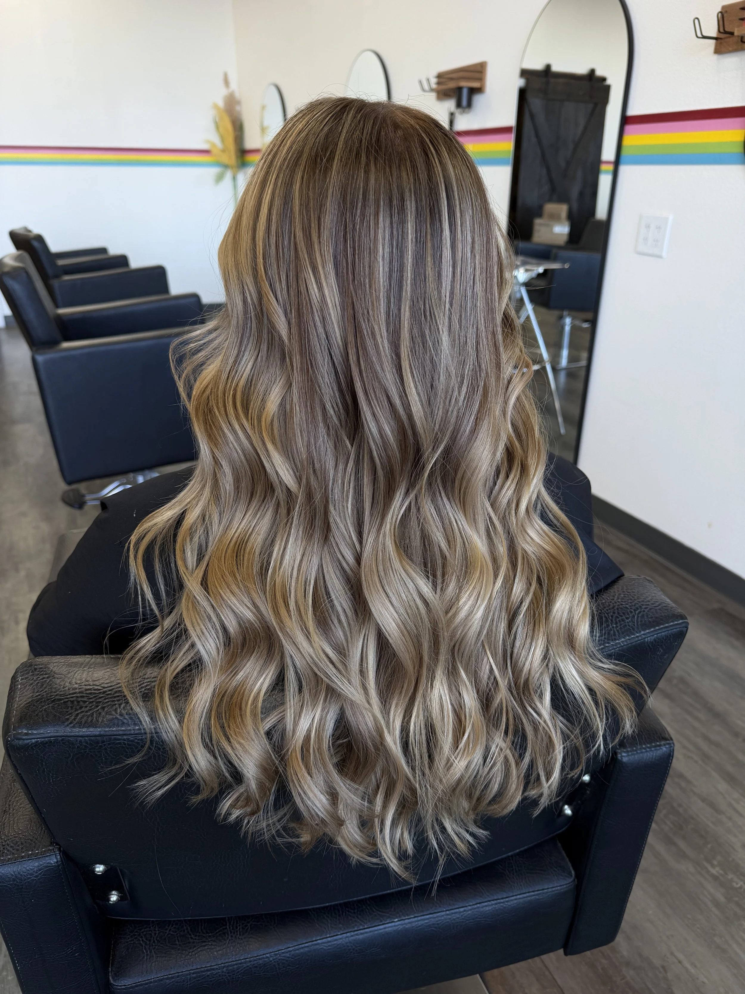 Back view of a woman with long wavy hair in shades of blonde and light brown, sitting in a salon chair.