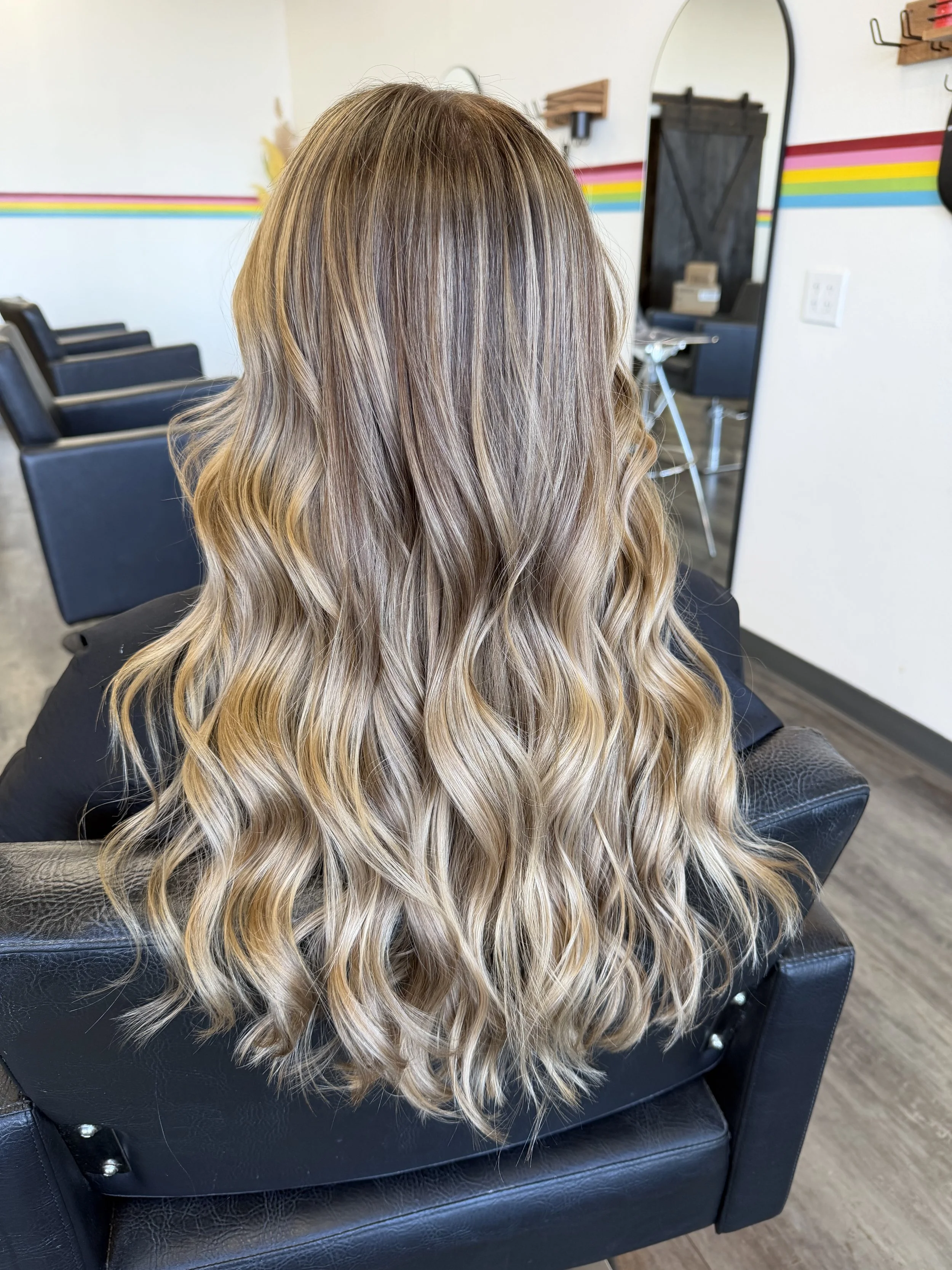 The image shows the back of a person with long wavy hair in shades of blonde and light brown, sitting in a black salon chair inside a hair salon.