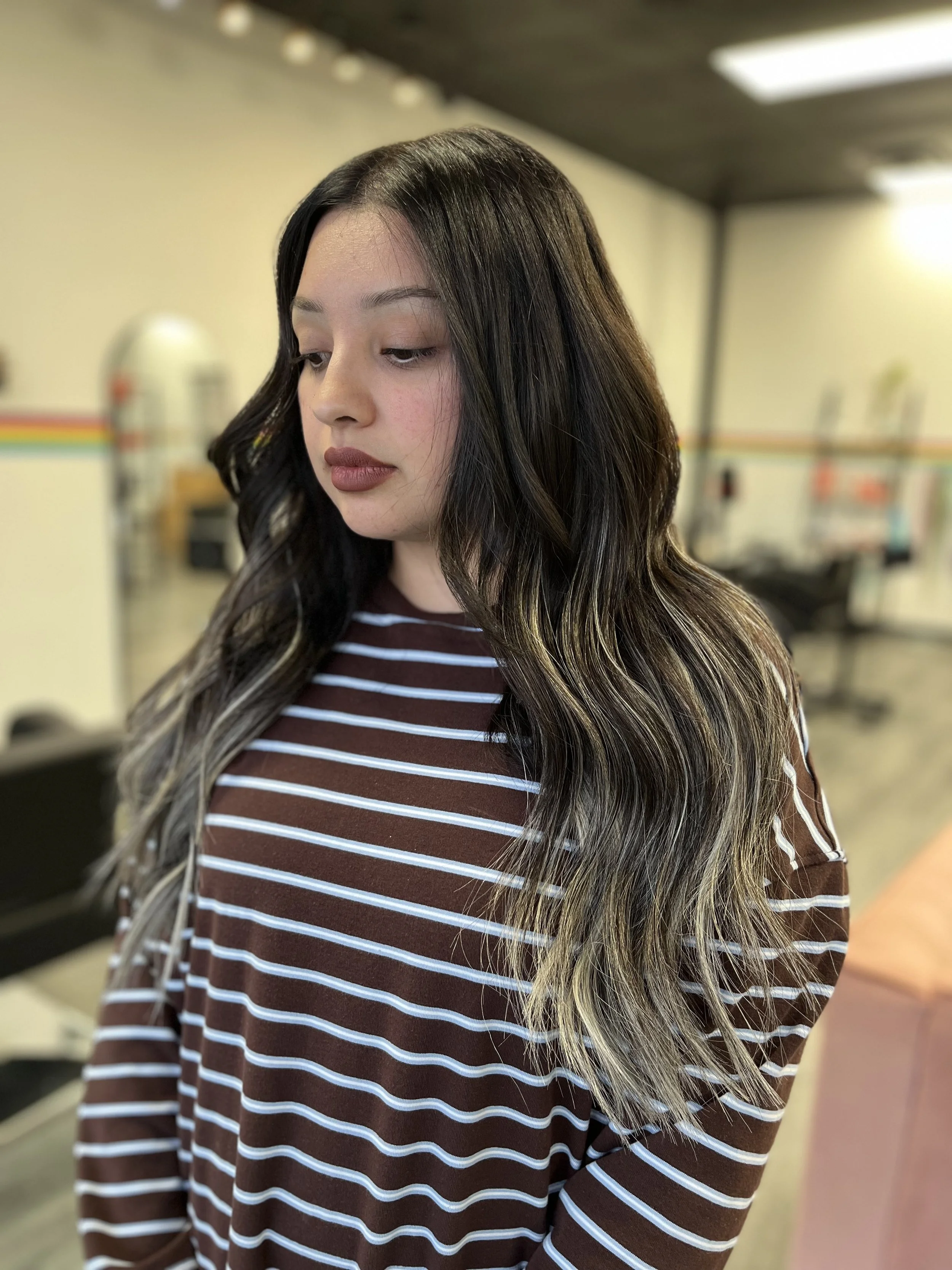 A woman with long dark hair and blonde highlights, wearing a brown and white striped shirt, is looking down with a neutral expression in an indoor setting.