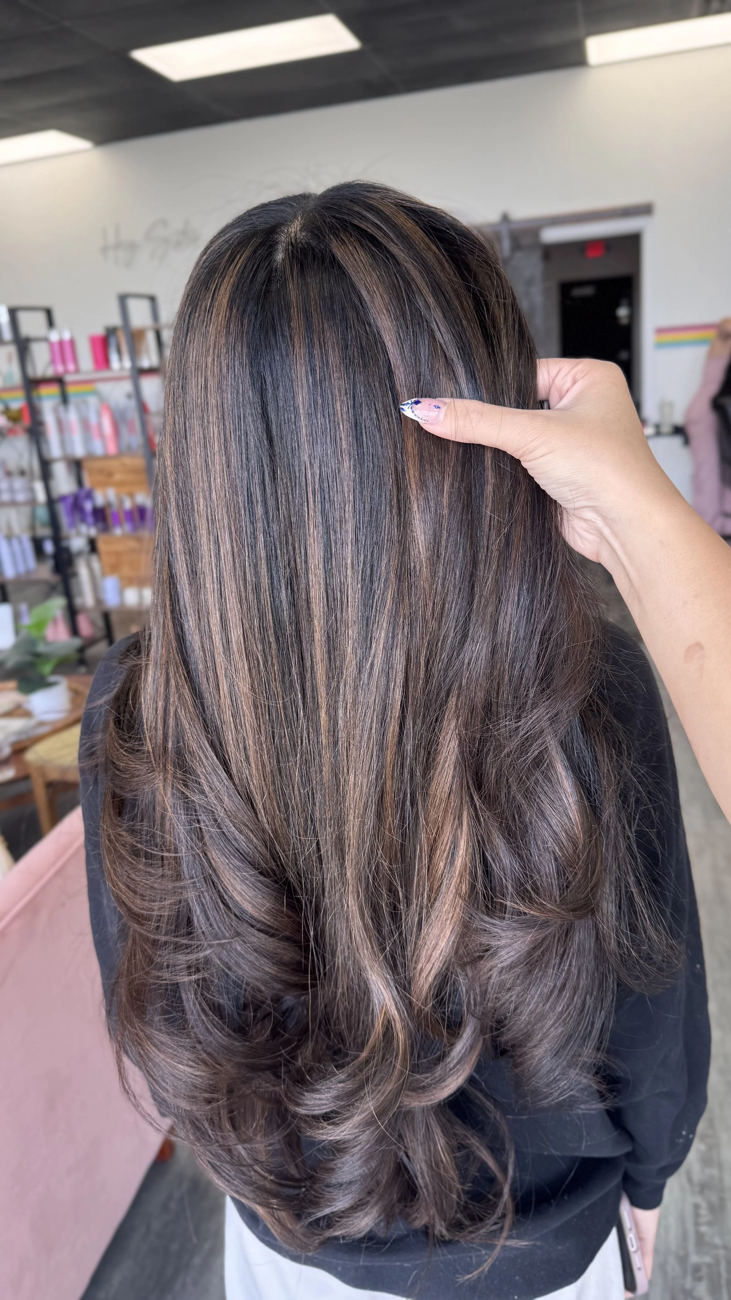 Back view of a woman with long, wavy hair that is dark at the roots and gradually lightens to shades of brown and blonde towards the ends, in a salon or studio setting.
