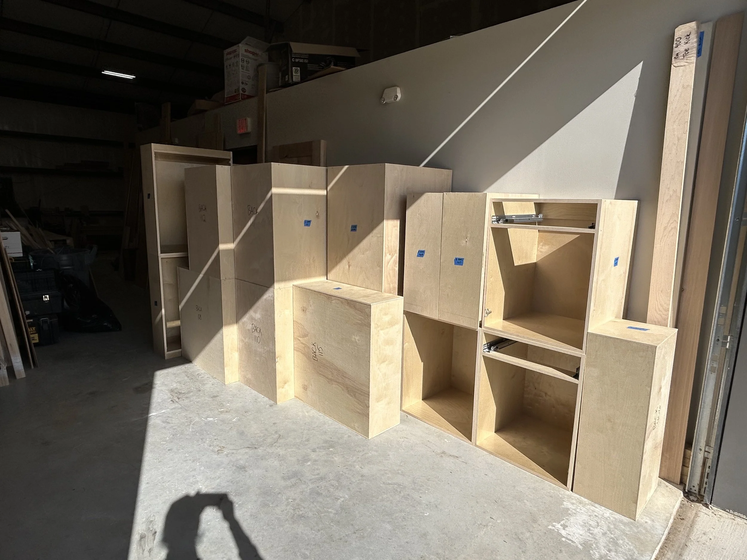 Unfinished wooden cabinets and shelves lined up in a workshop, with sunlight casting shadows on the floor.