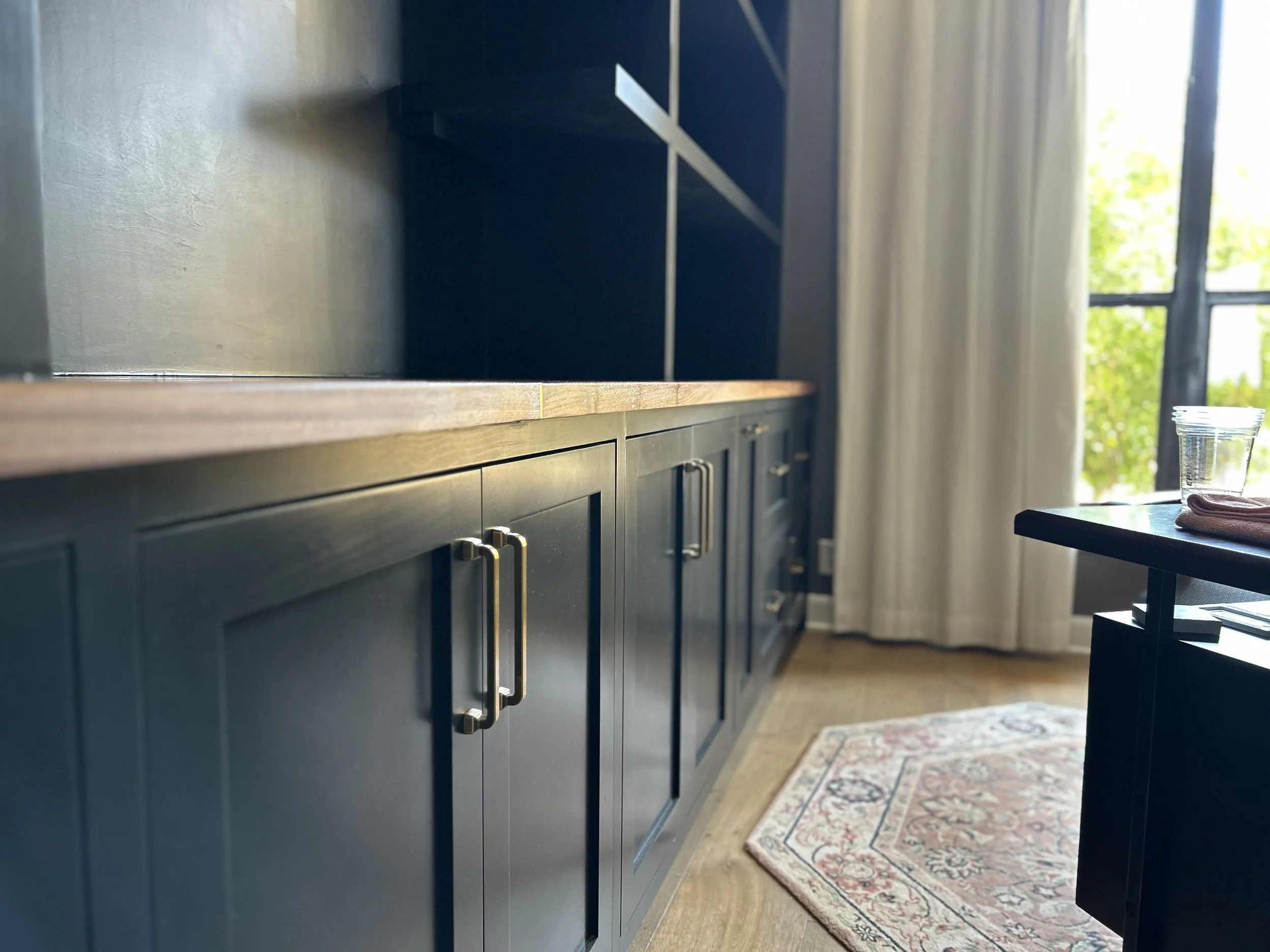Interior view of a kitchen with dark blue cabinetry, a wooden countertop, a window with white curtains, and a patterned rug on a hardwood floor. A glass and a cloth are on a small side table.