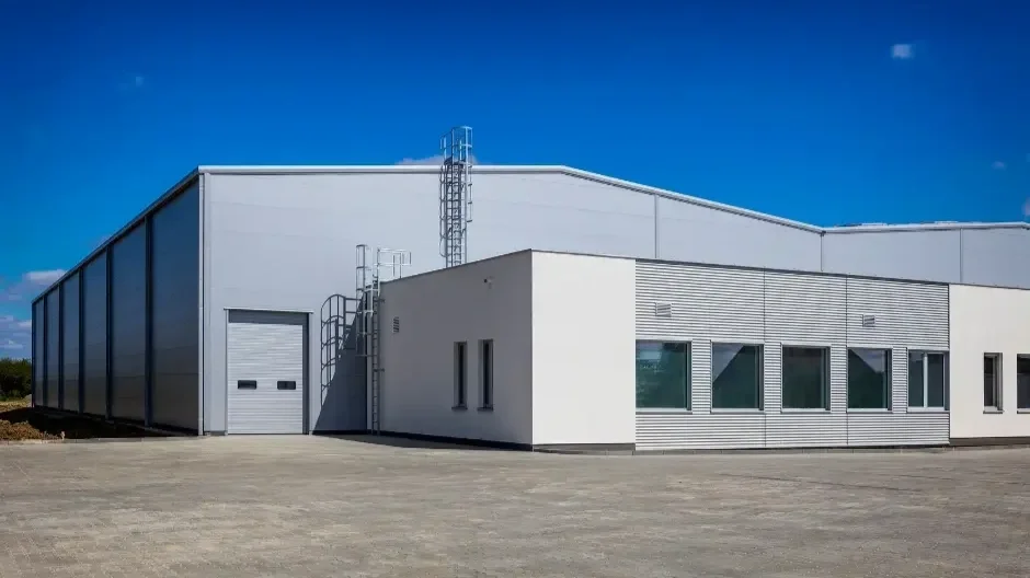 A large industrial warehouse building with a white exterior, a roll-up door, and several windows under a blue sky.