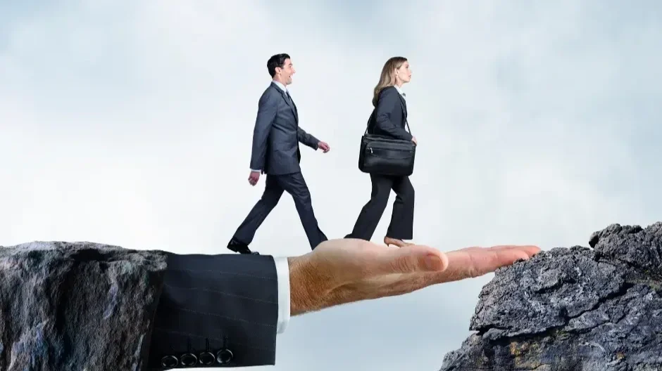 Two business people walking on a large hand, with a rocky ledge on either side, symbolizing taking a risk or support in a business environment.