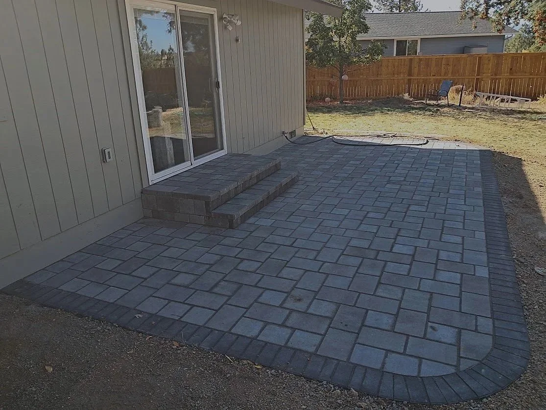 Newly constructed gray brick patio with steps leading to sliding glass door, surrounded by a wooden fence and backyard with grass, trees, and outdoor furniture.