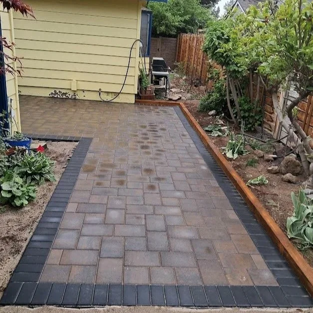 Backyard paver patio, bordered by black pavers, beside a garden bed with plants, a yellow house wall, and a wood fence.