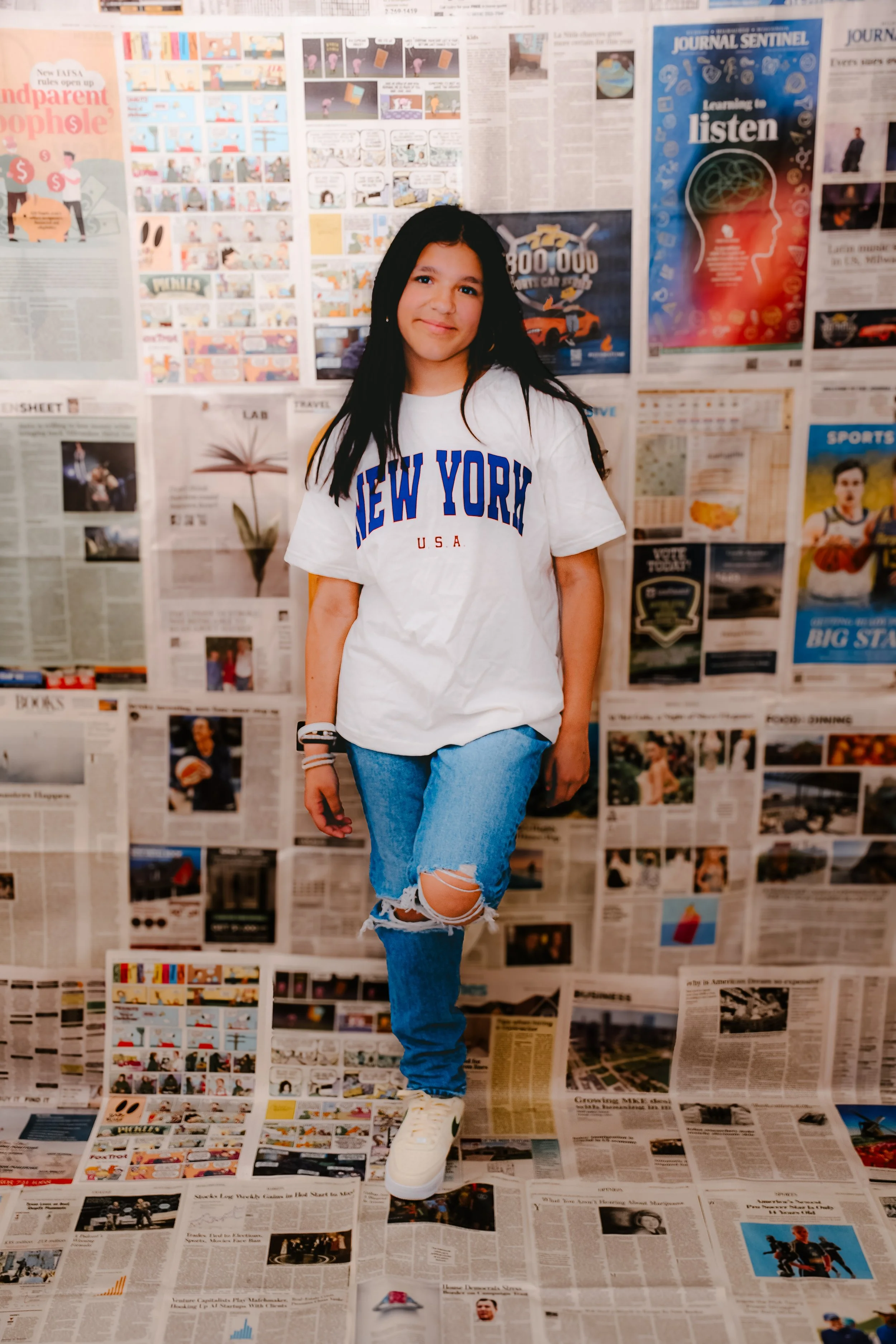 A young girl with long dark hair wearing a white t-shirt with blue and red text saying 'NEW YORK U.S.A.', ripped blue jeans, white sneakers, stands in front of a wall covered in newspaper pages, with some newspapers on the floor.