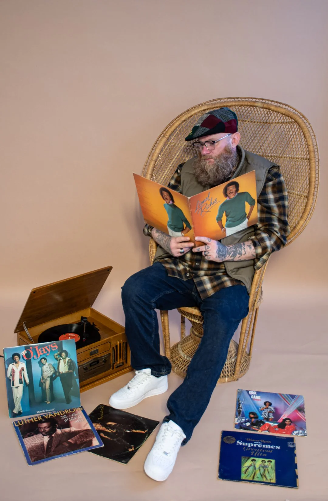 A man with glasses, a beard, tattoos, and a checkered shirt, wearing a green vest and a plaid hat, sits on a wicker chair reading a vinyl album cover. There are several vinyl albums on the floor and next to a vintage record player.