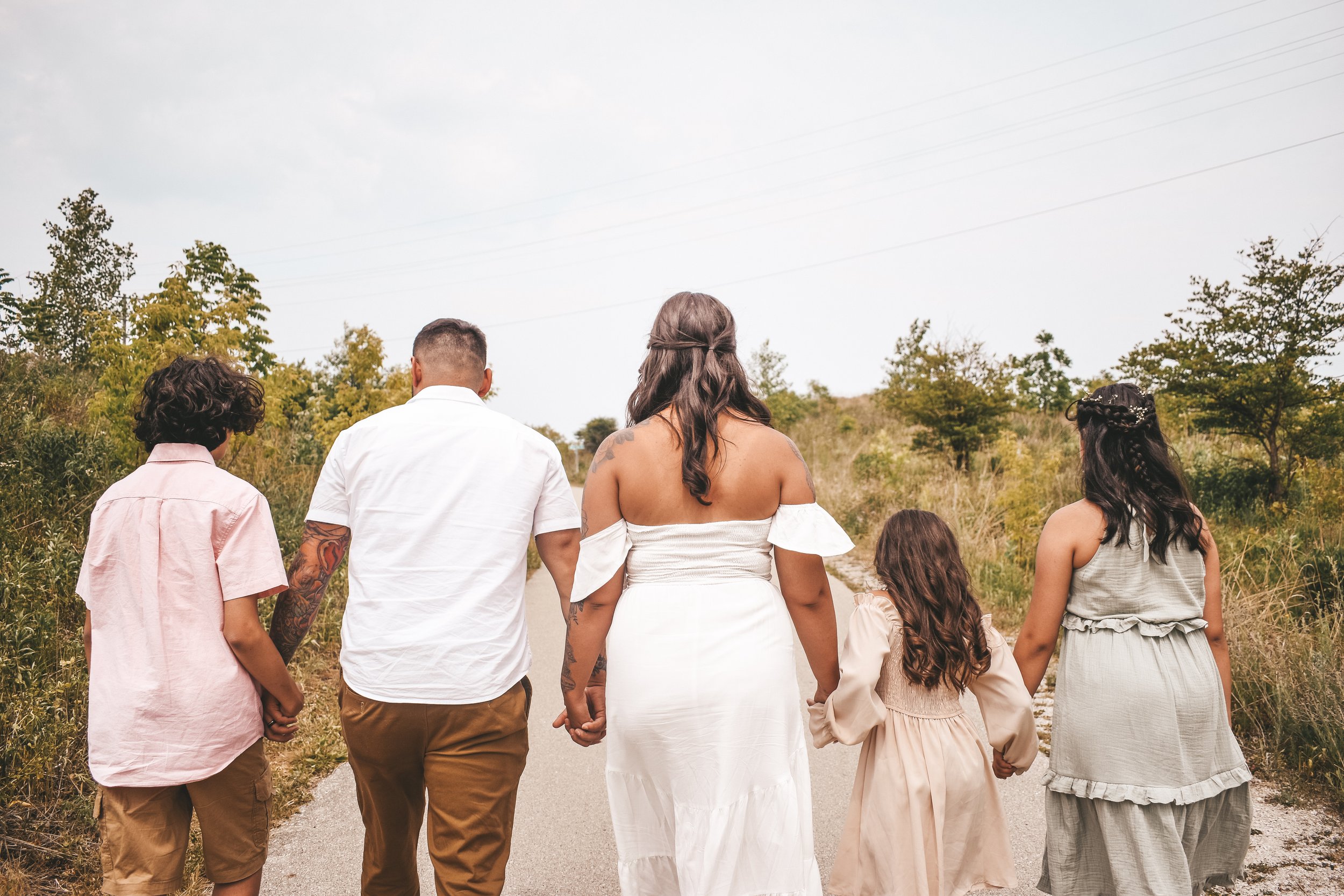 Family of five holding hands and walking down a rural path surrounded by greenery and trees.