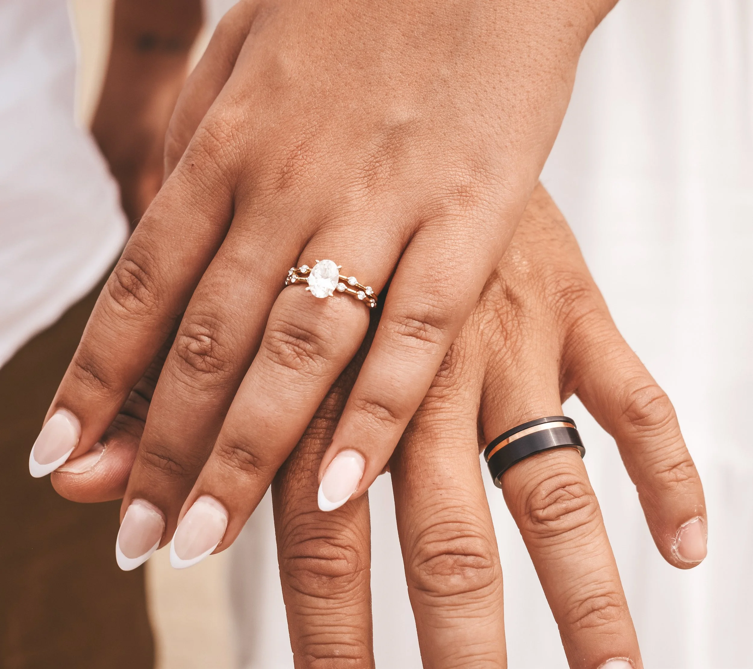 Two hands with wedding rings, one with an oval diamond ring and the other with a black and gold band, overlapping.