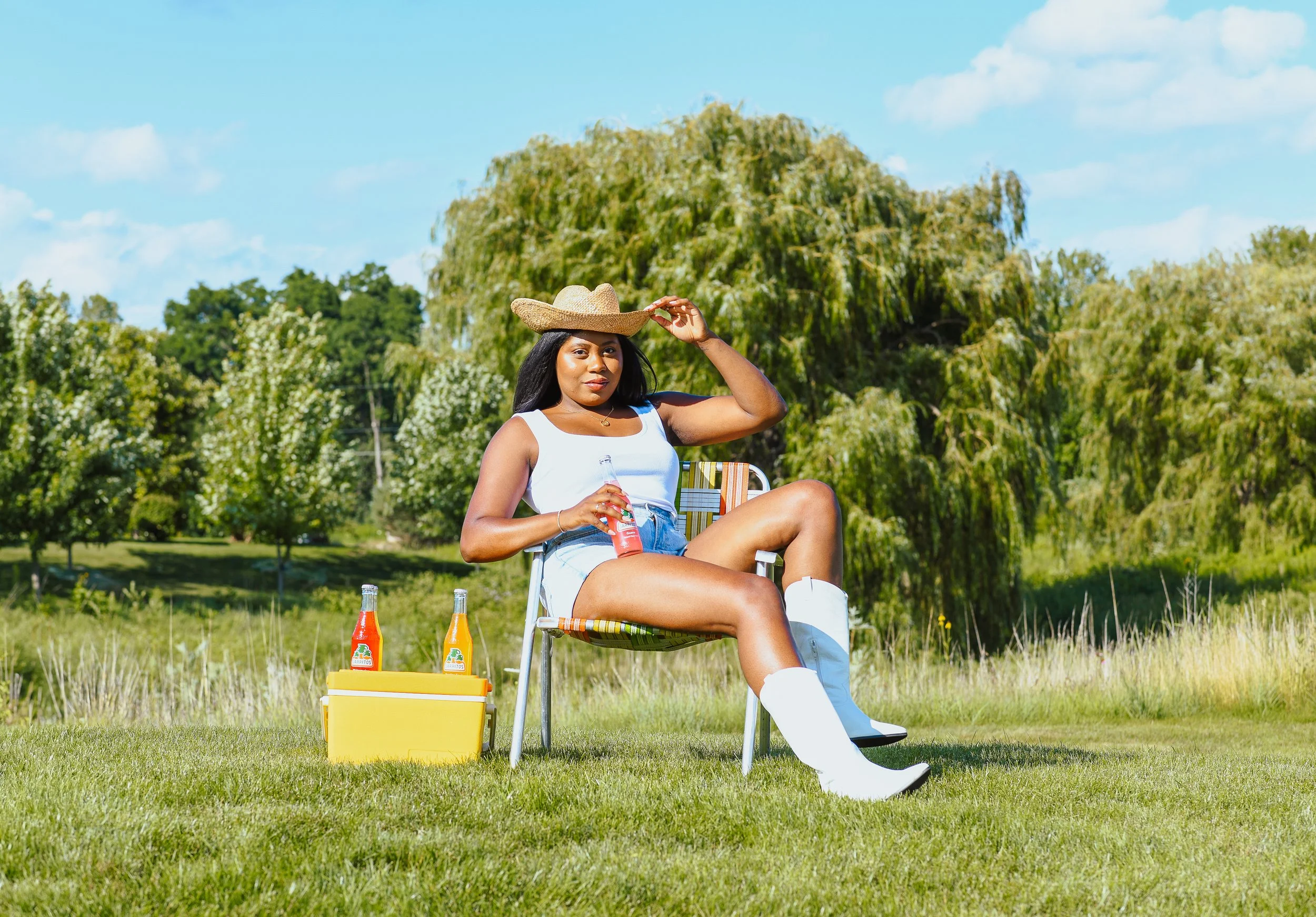 Woman sitting outdoors on a lawn with a yellow cooler and two bottles of soda, holding a soda, wearing a white tank top, denim shorts, white cowboy boots, and a large straw hat, with trees in the background on a sunny day.