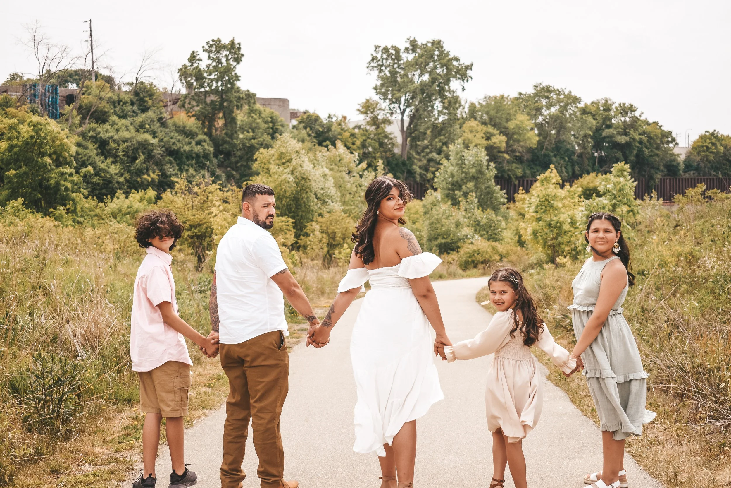 A family of six holding hands and standing on a nature trail, with trees and greenery in the background, during daytime.