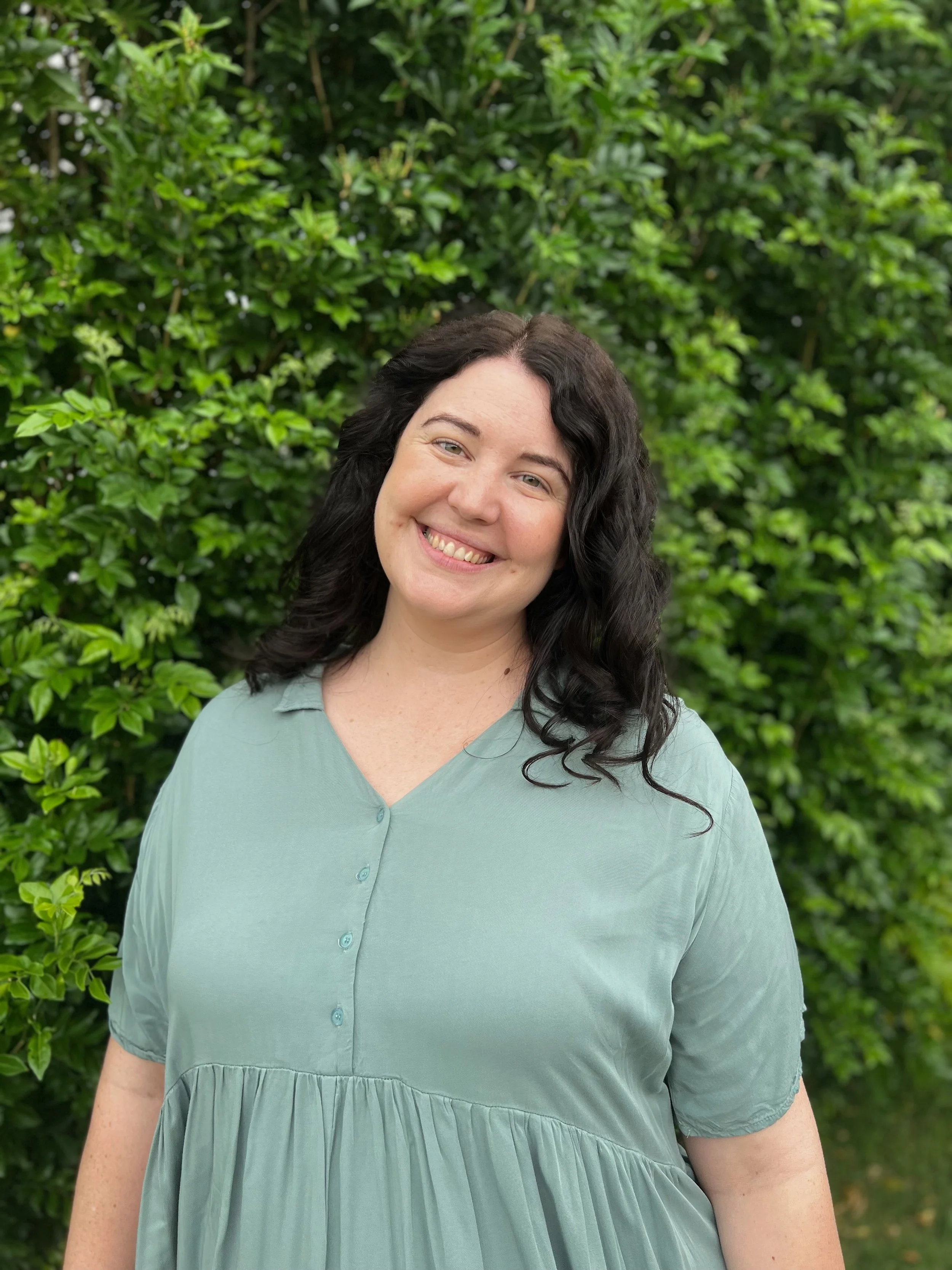 A woman smiling outdoors with green foliage in the background, wearing a light green dress.