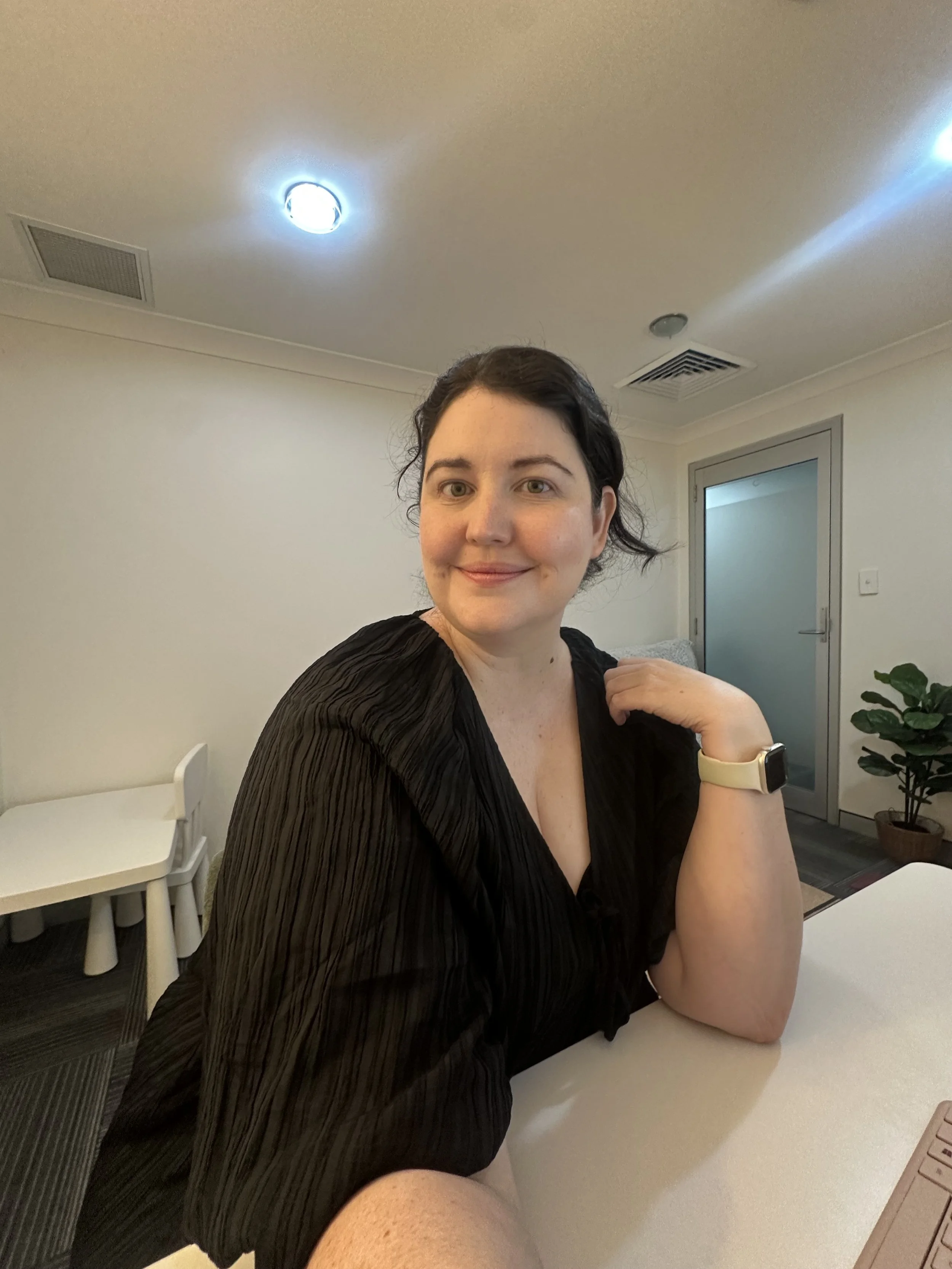 A woman with dark hair, wearing a black top and a yellow smartwatch, sitting at a table in a modern room with white walls, a door, a houseplant, and a stack of white chairs.