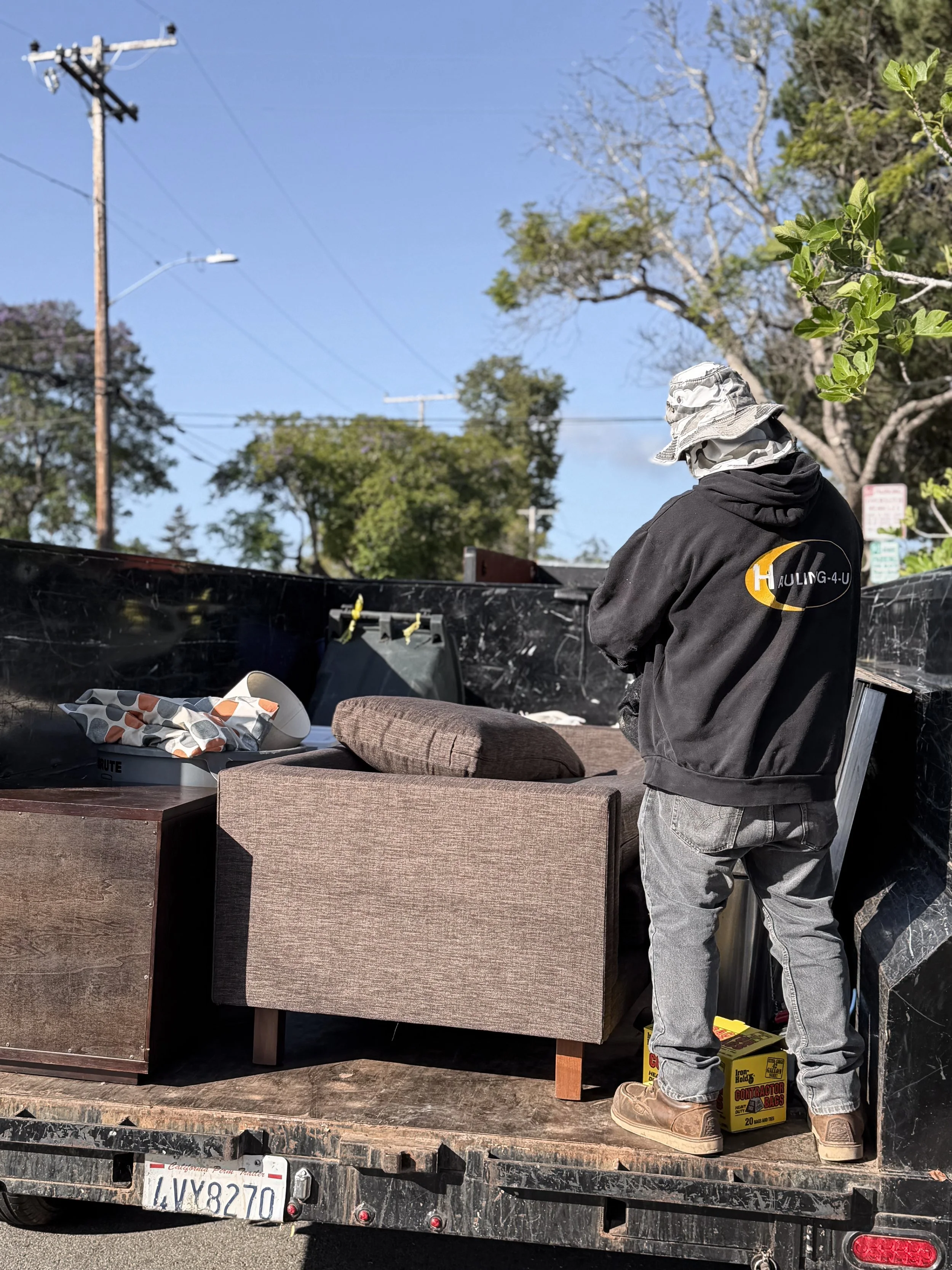 A person wearing a camo bucket hat, black hoodie with a logo, and gray pants is standing at the back of a truck, looking away from the camera, with furniture and miscellaneous items loaded on the truck bed, including a box of contractor bags, a beige couch, and a table. There are trees and power lines visible in the background.
