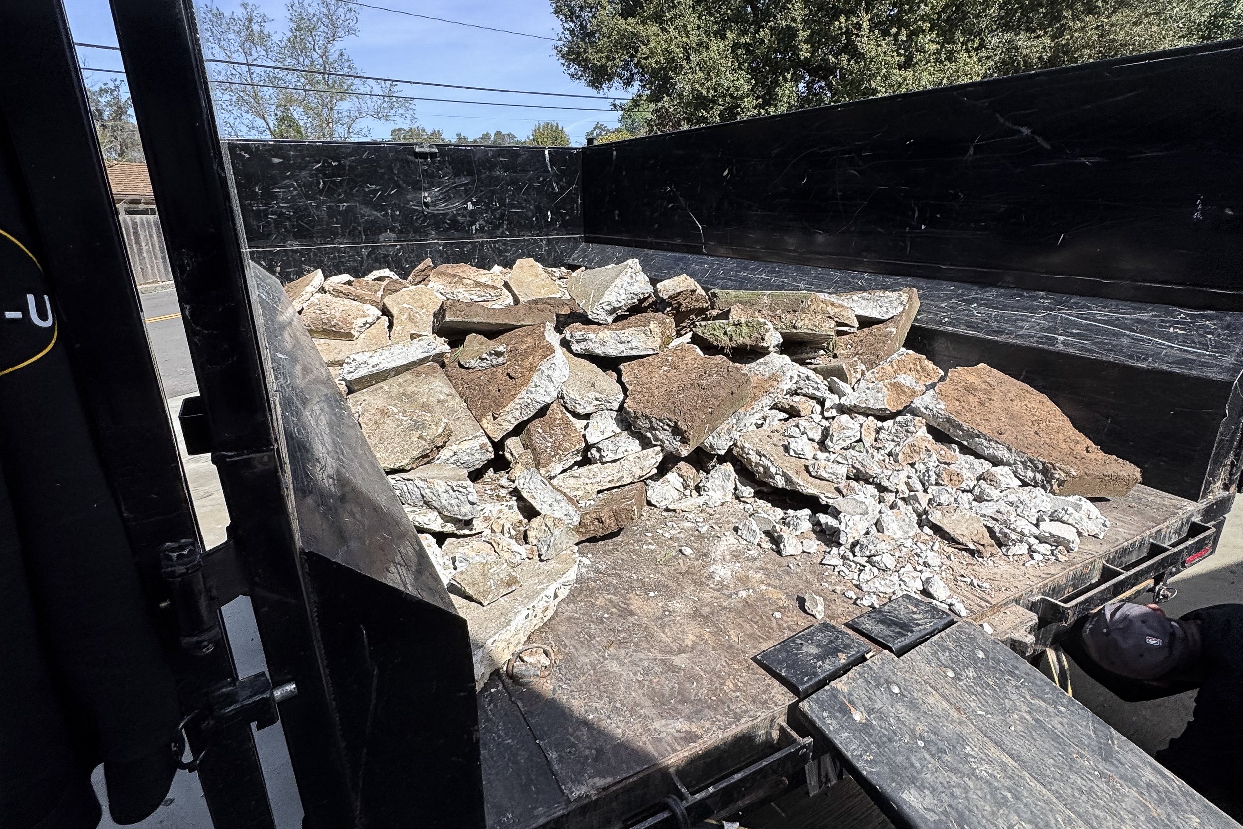 The image shows the inside of a black dump truck bed filled with various sizes of rocks and stones, parked outdoors under a clear sky.