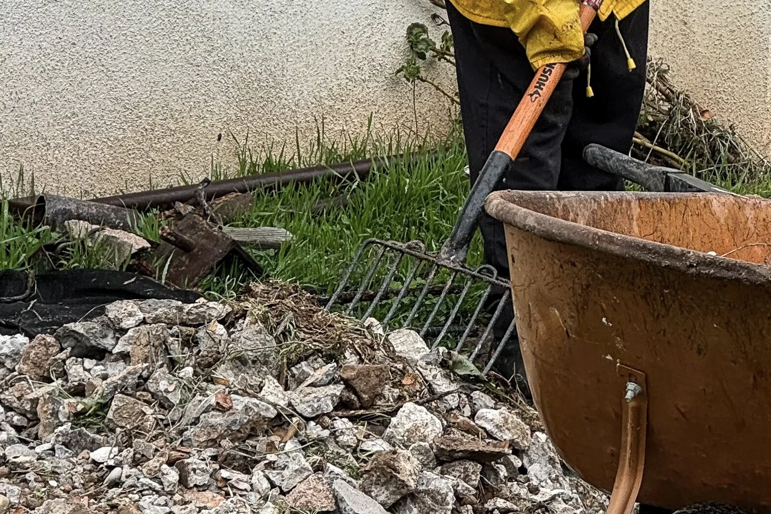A person working on a construction or landscaping project outdoors, with a wheelbarrow, a rake, and debris including rocks and metal tools near a building wall.
