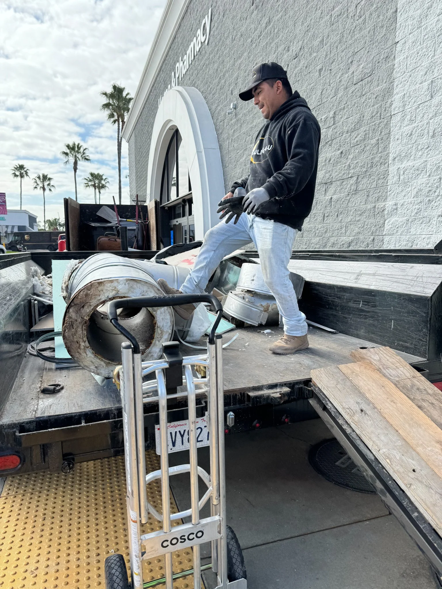 A man standing on a flatbed truck loading an air conditioning unit, with a dolly nearby, outside a modern building with palm trees in the background.