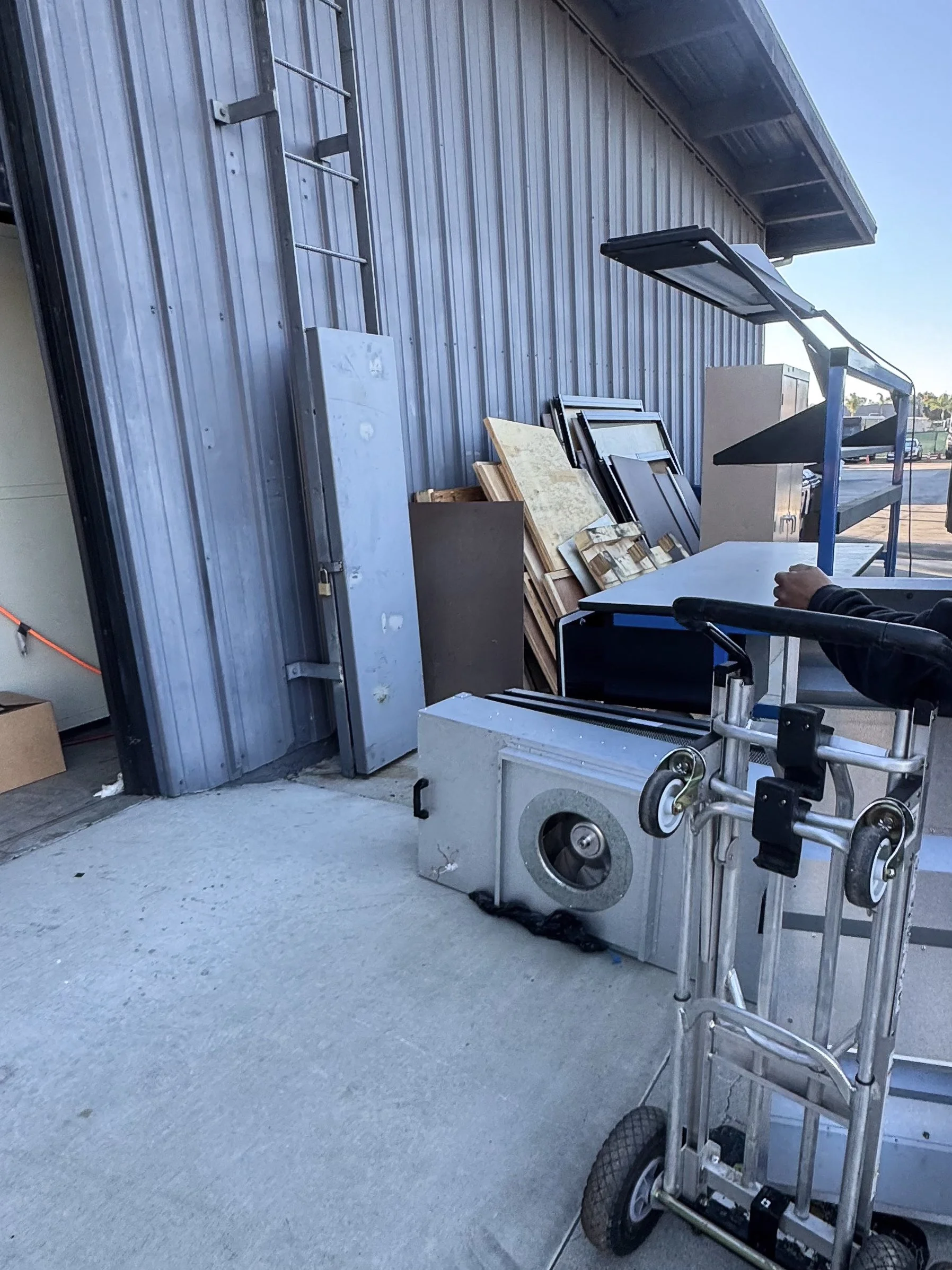 Outdoor scene of a building with metal siding and a garage door. A ladder, some wood planks, and a metal cart are visible, along with a person operating a wheelchair.