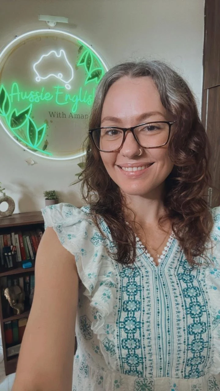 A woman with curly brown hair, glasses, and a nose piercing smiling for a selfie in front of a neon sign that says 'Aussie English with Amanda' with a kangaroo outline and green leaves. She is wearing a white and blue embroidered top and standing in a room with a bookshelf and small potted plants.