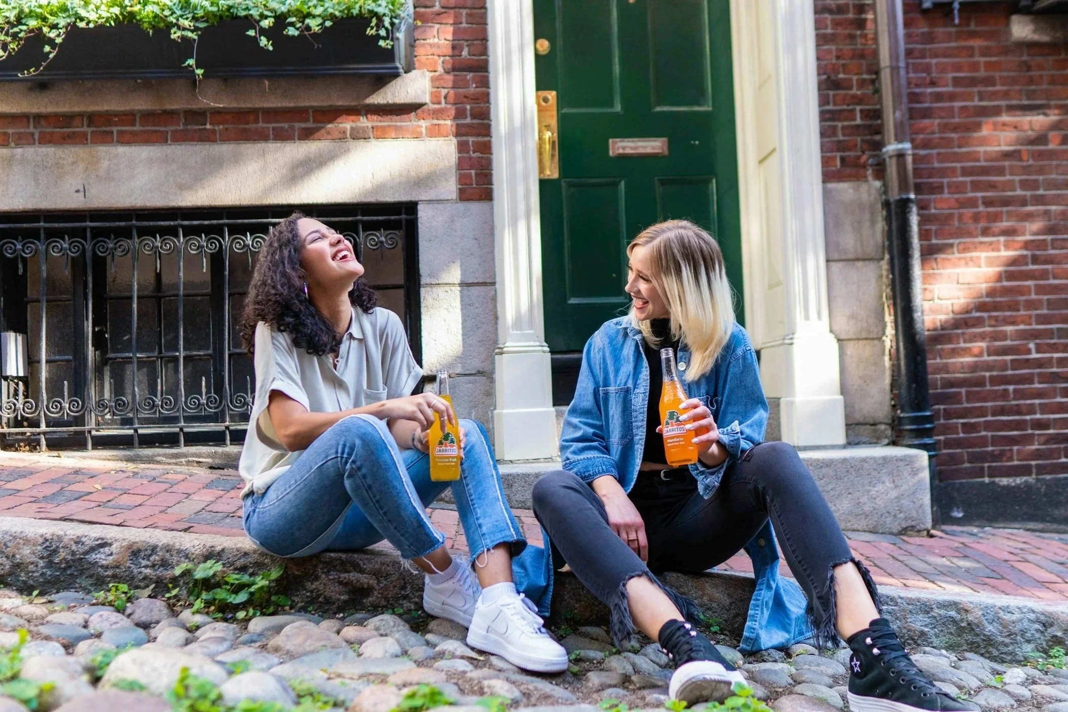 Two young women sitting on a cobblestone sidewalk, laughing and enjoying drinks in bottles, in front of a brick building with a green door.