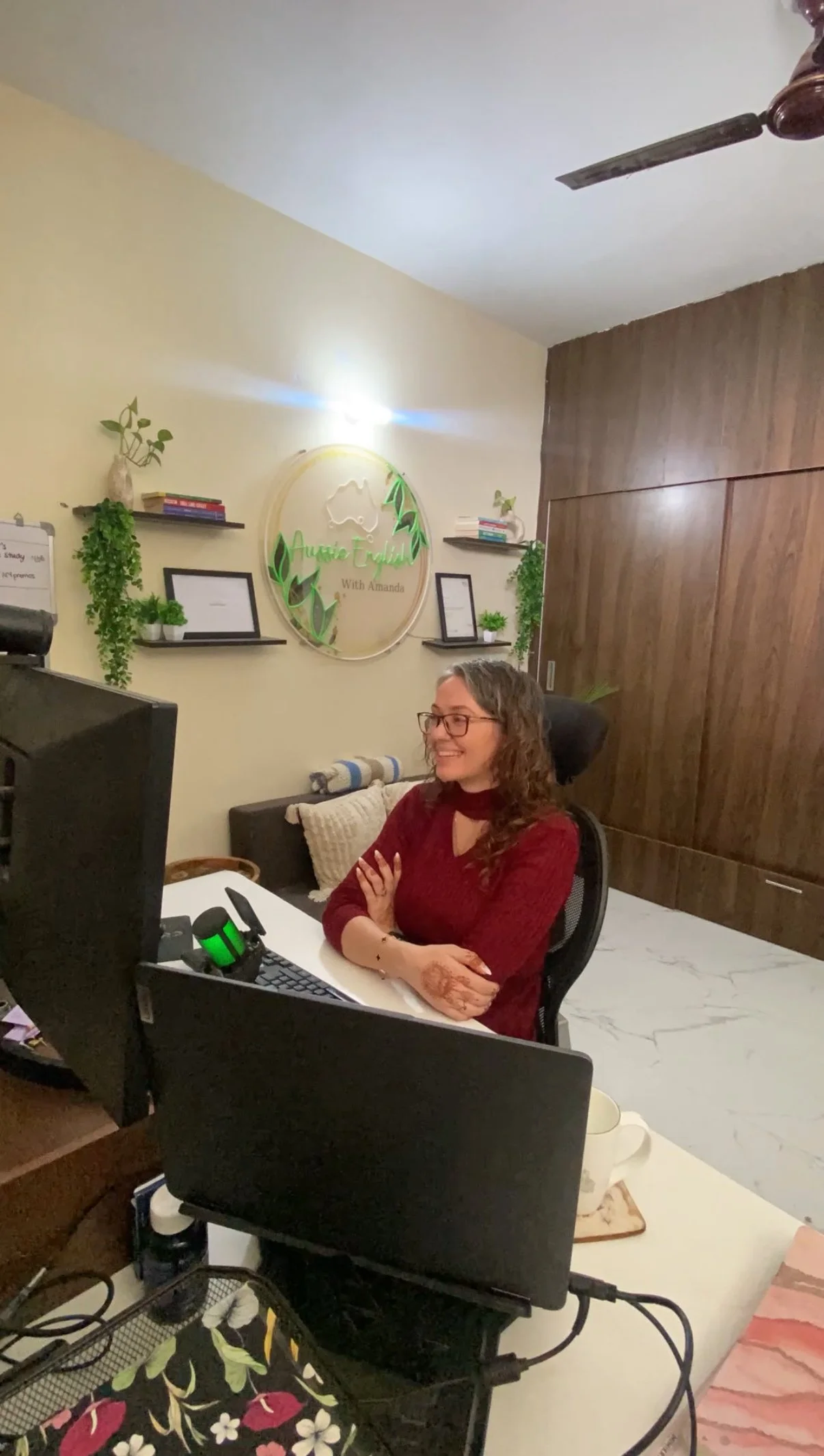 A woman with glasses and long curly hair, smiling, sitting at a desk with a computer monitor, keyboard, mouse, and headphones in an office or home workspace. The wall behind her has a decorative sign that says 'Aussie Travails with Amanda,' framed pictures, and hanging plants.