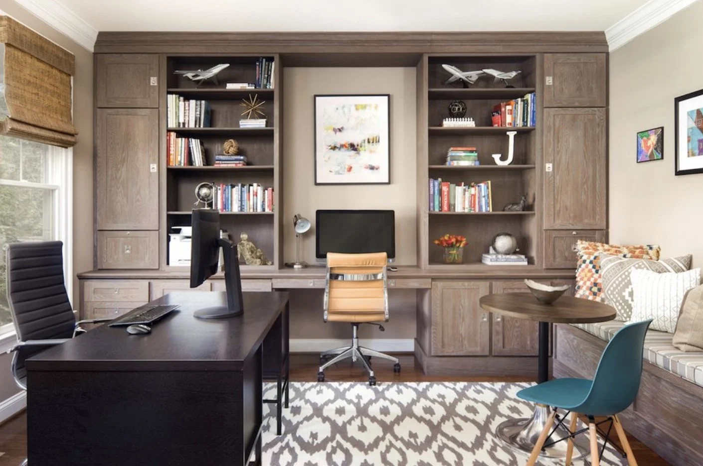 Home office with black desk, computer, beige desk chair, built-in wooden shelving with books, decorative items, and planes, gray patterned rug, window with blinds, and seating area with colorful pillows.