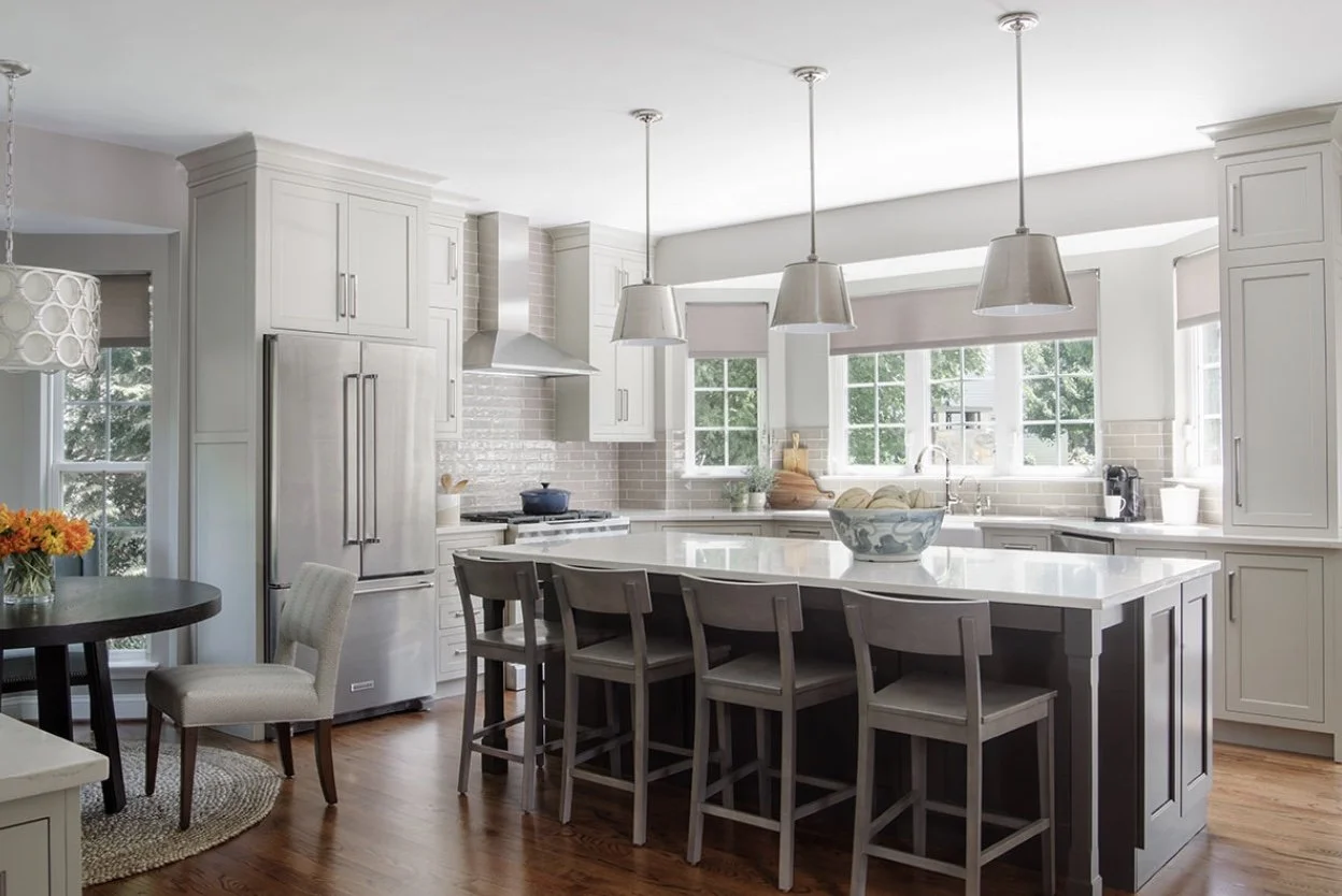 Bright modern kitchen with white cabinets, a large island with a dark base and white countertop, stainless steel appliances, and wooden flooring. Sunlight streams through multiple windows.