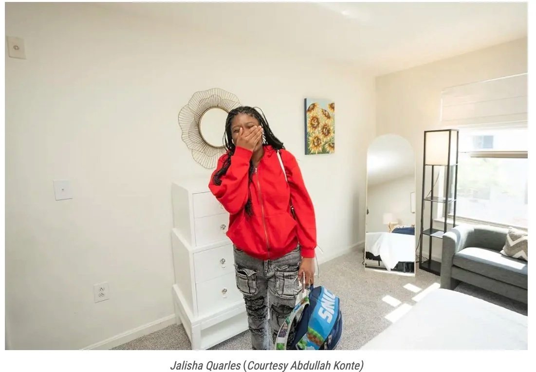 An African American woman in jeans and a red hoodie covering her mouth an laughing in disbelief in her new bedroom.