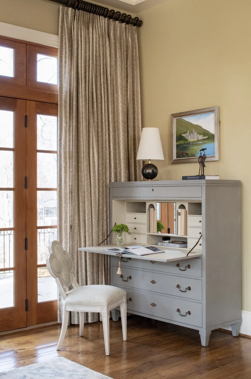 Asymmetrical balance styled on a vintage secretary desk with a designer lamp, artwork, and books for a high-end home office look.