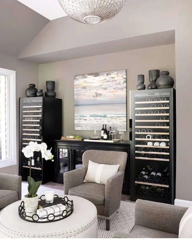 A living room with two wine storage cabinets, a beige armchair with a pillow, a round white ottoman with a tray, a plant, and a painting of the ocean on the wall.