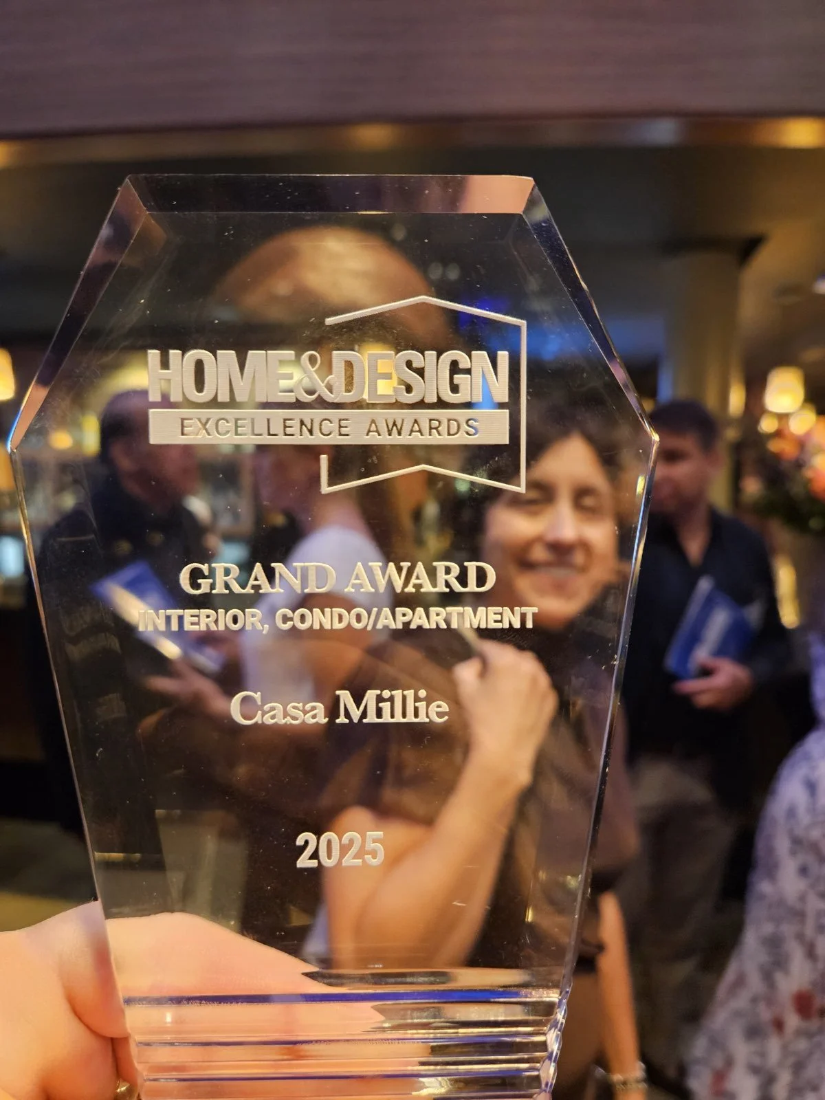 A clear trophy with gold inscription, held in front of a smiling woman and others at an awards event, reading 'HOME & DESIGN EXCELLENCE AWARDS GRAND AWARD INTERIOR, CONDO/APARTMENT Casa Millie 2025'.