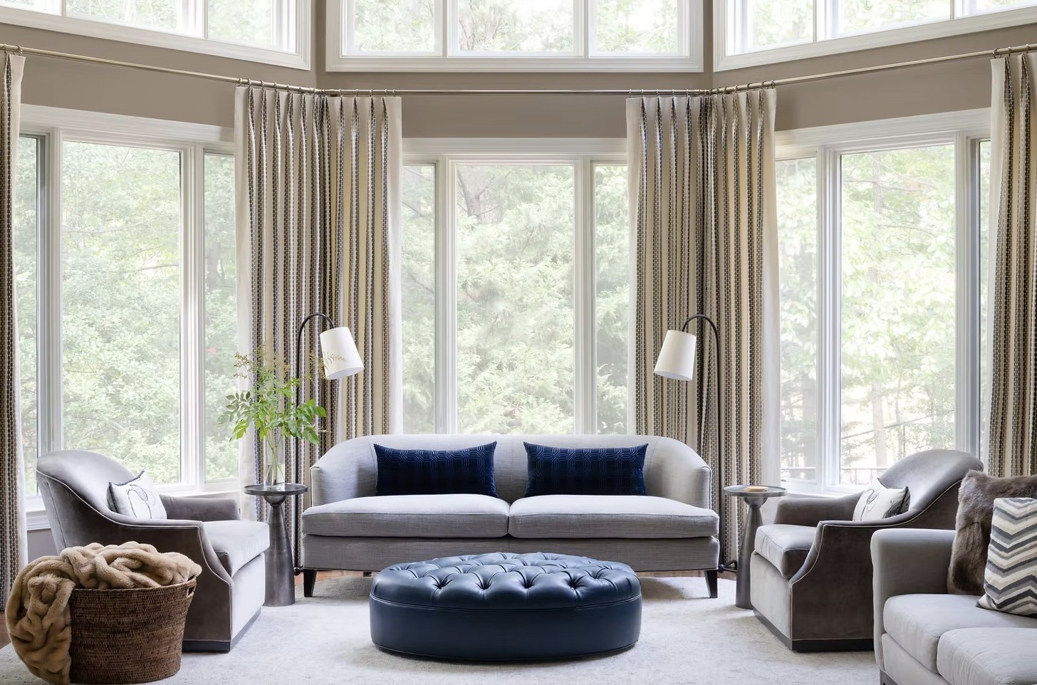 Living room with large bay windows, beige curtains, grey sofas with blue pillows, two armchairs, a black ottoman, lamps, a plant in a basket, and decorative pillows.