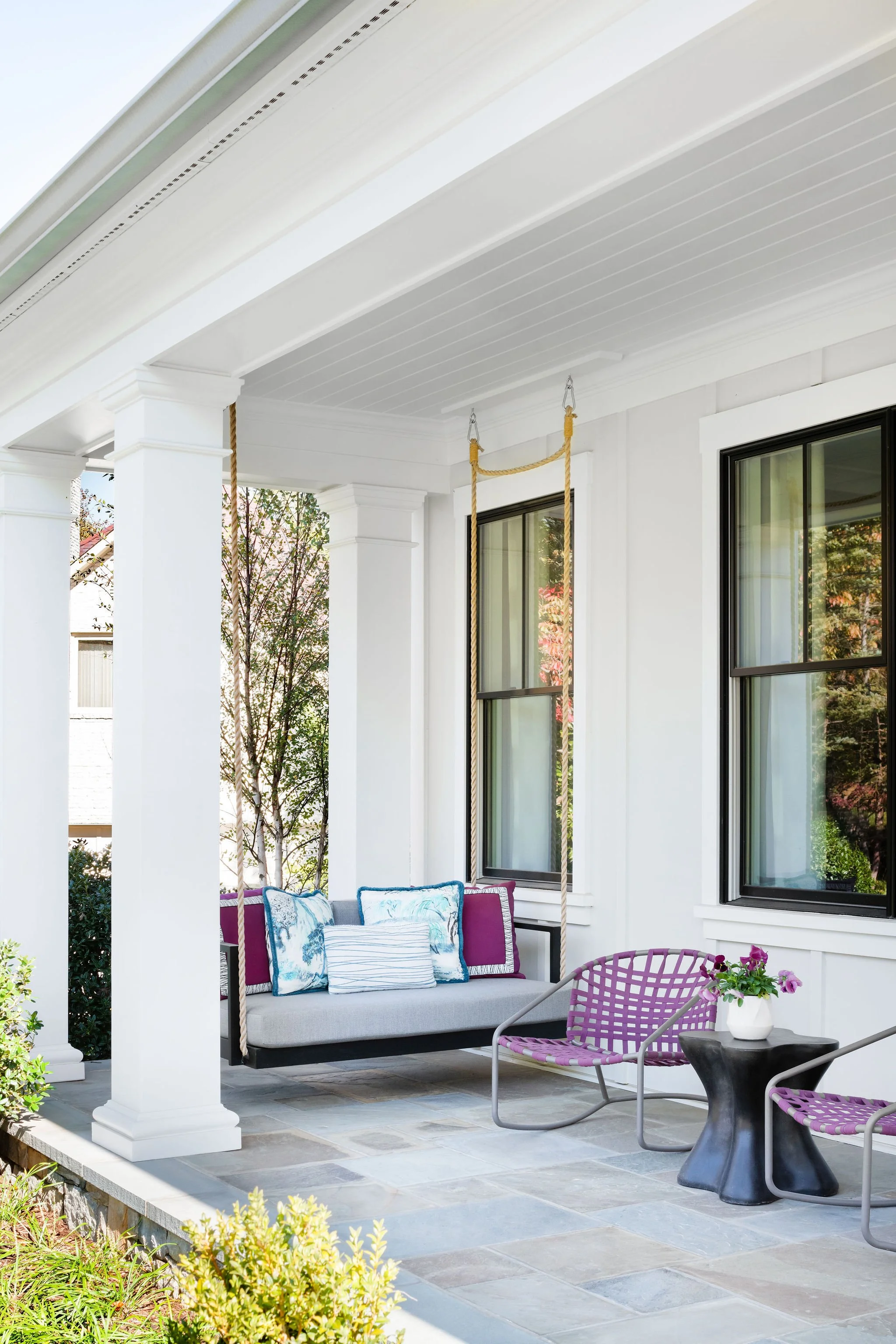I white columned front porch of a Vienna, Virginia home with a gray porch swing and purple patio chairs.