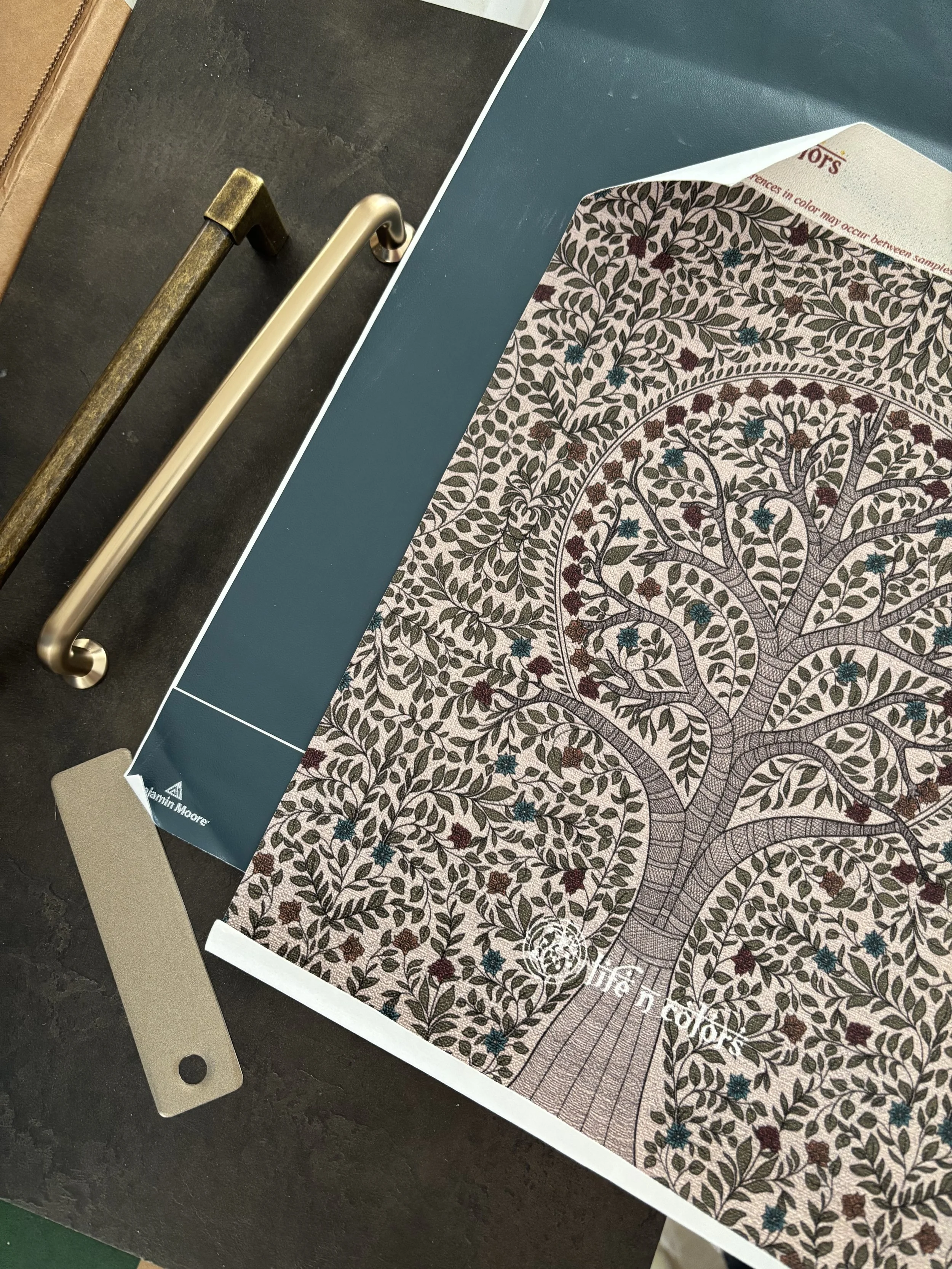 Polished brass drawer pull held against warm-toned Madhubani botanical wallpaper for material selection.