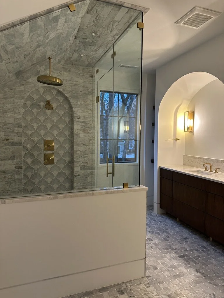 Luxury primary bathroom with wood vanities, marble surfaces, and an arched shower entrance.