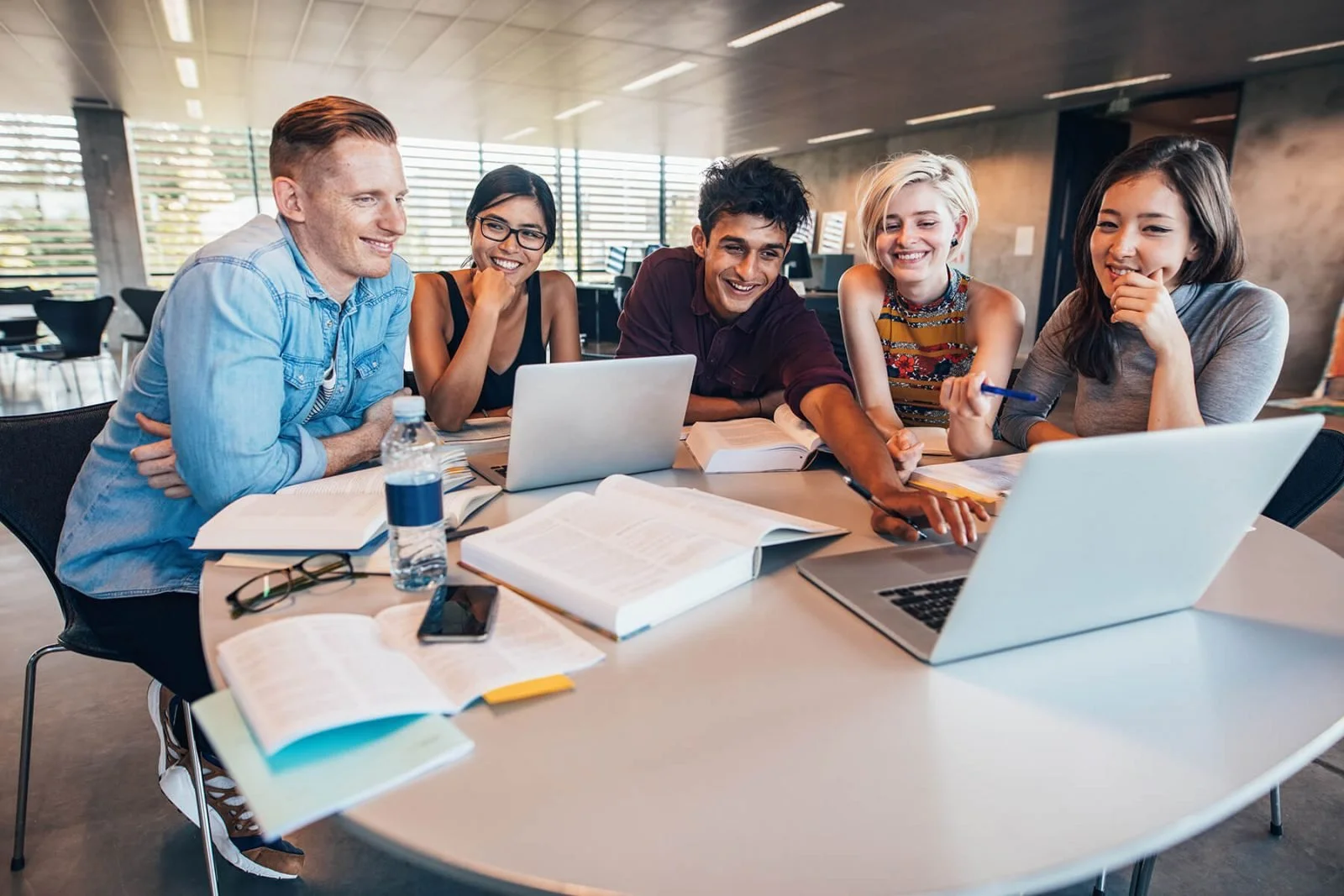 Group of five young adults sitting around a table, smiling and looking at a laptop in a modern office space with books, notebooks, and a water bottle on the table.