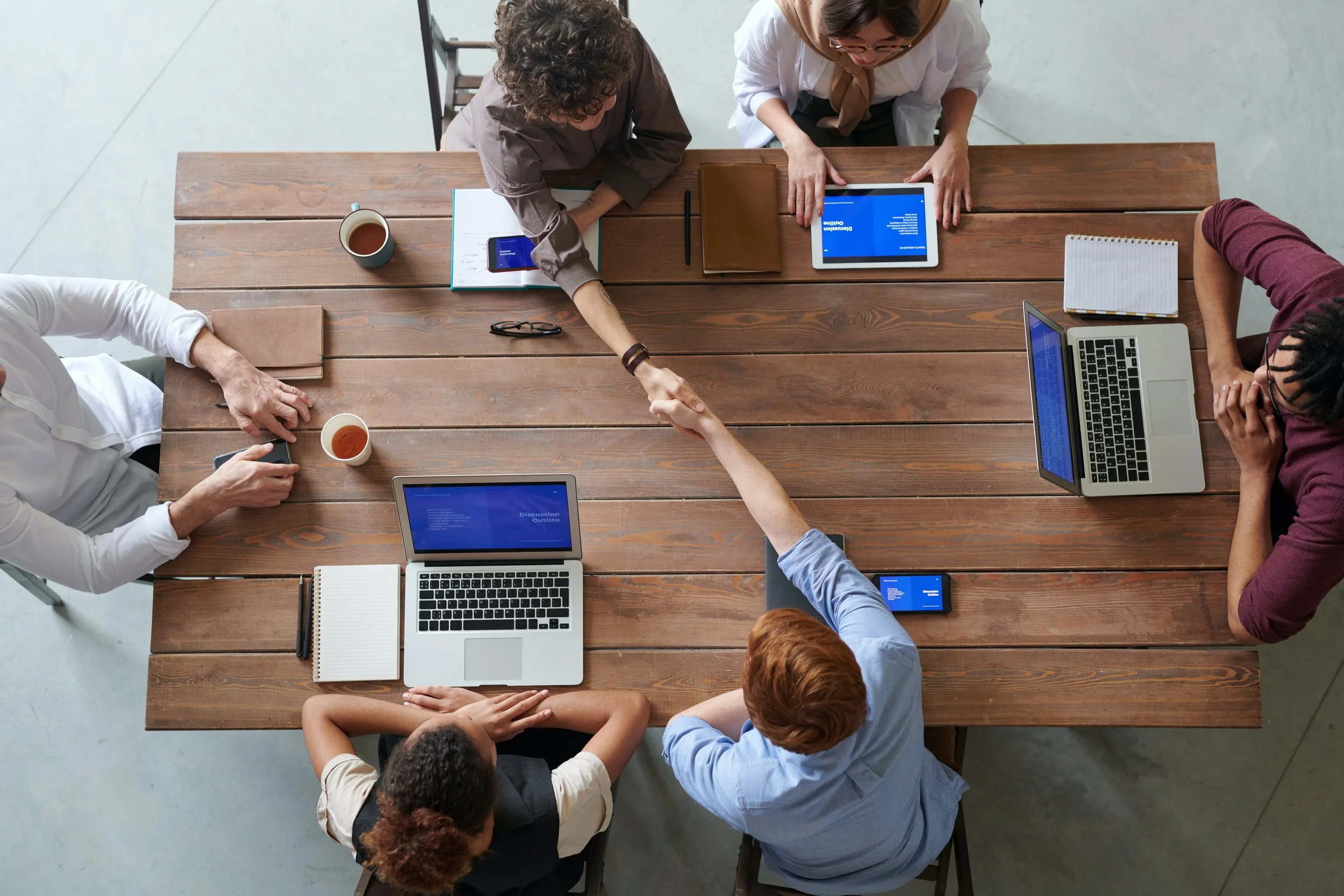 Six people sitting around a wooden table engaged in a business meeting, shaking hands, with laptops, notebooks, and cups of coffee on the table.