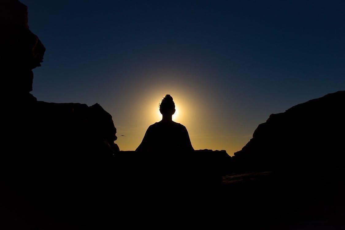 Silhouette of a person sitting between two rock formations during sunset or sunrise with the sun behind them.