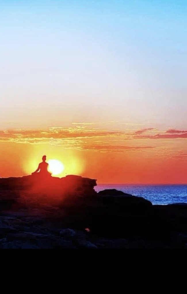 Silhouette of a person meditating on rocks by the ocean during sunset, with the sun setting behind them and colorful sky overhead.