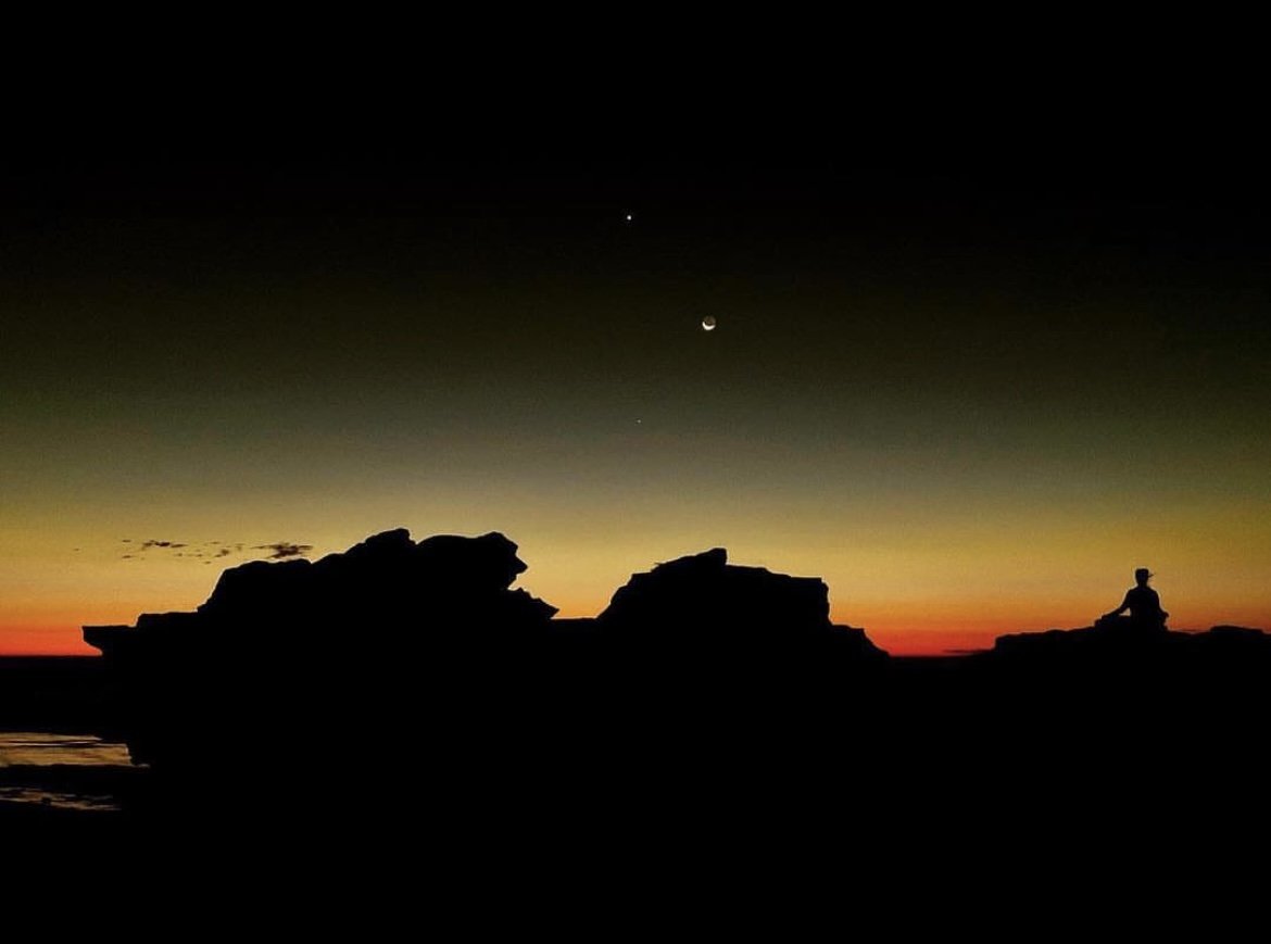 Silhouette of a person meditating on rocks during sunset with a colorful sky featuring a bright star, a crescent moon, and a distant planet.