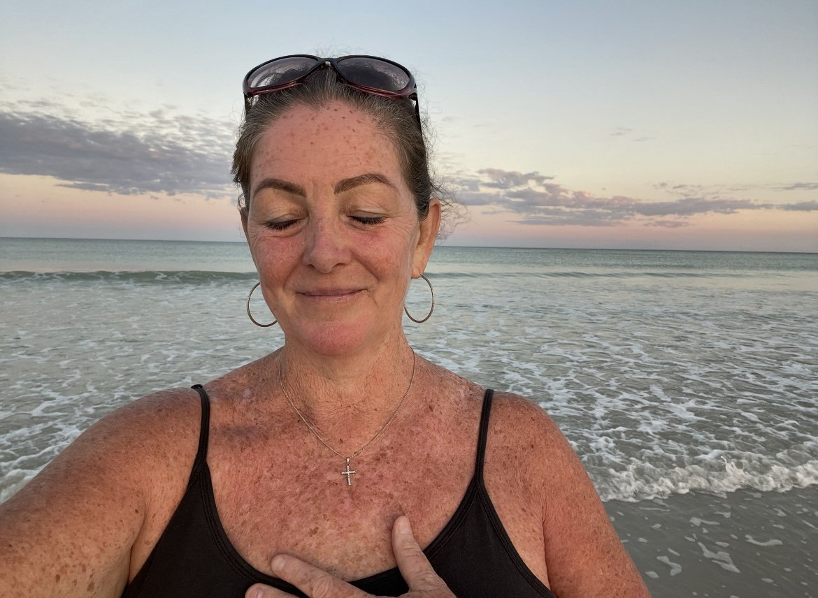 A woman with freckles and sunglasses on her head at the beach, eyes closed, smiling softly, wearing a black top, necklace with a cross, and hoop earrings, during sunset or sunrise.